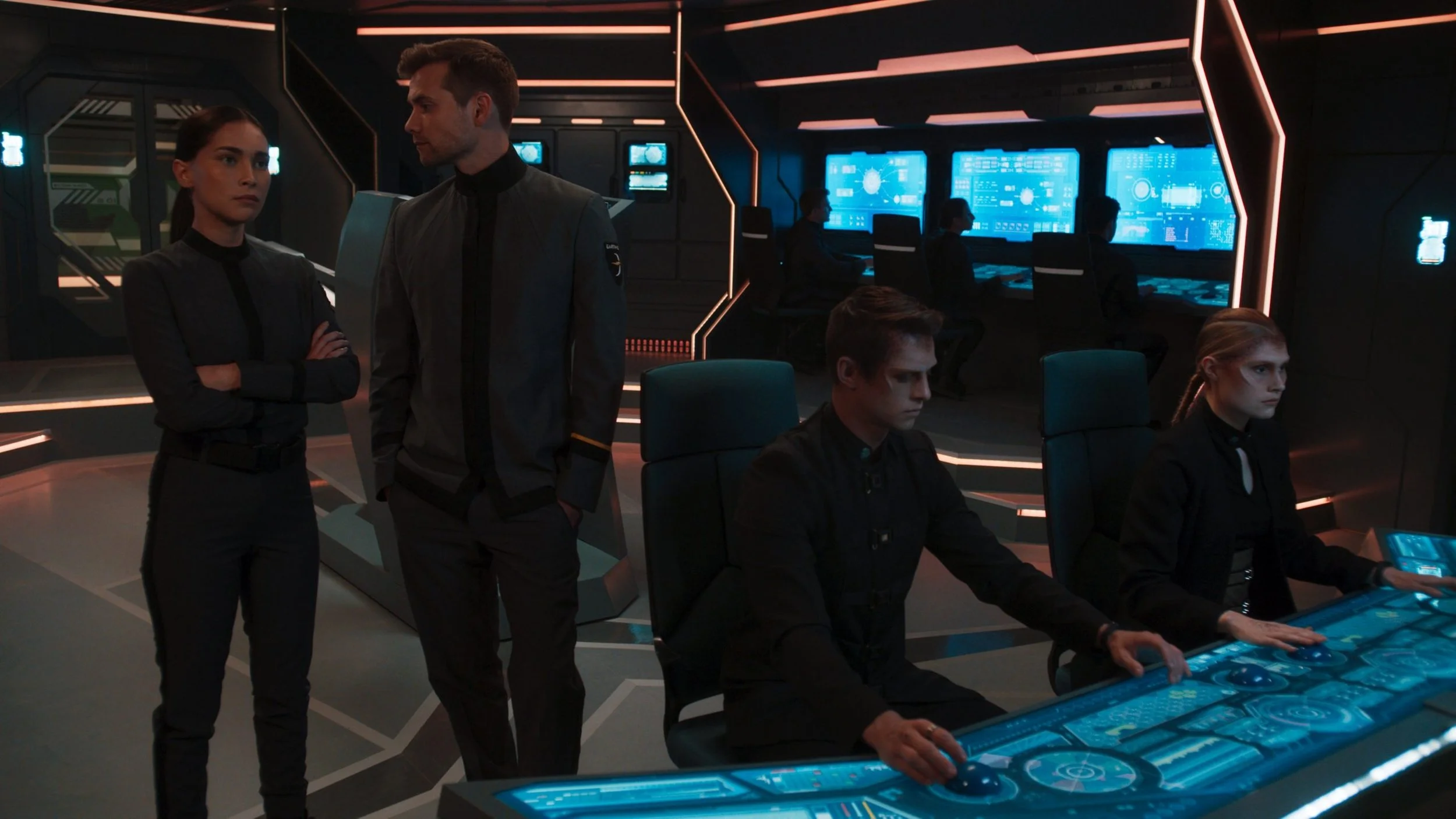 A group of people in a futuristic control room with large blue digital screens and consoles, some working and others standing and observing.