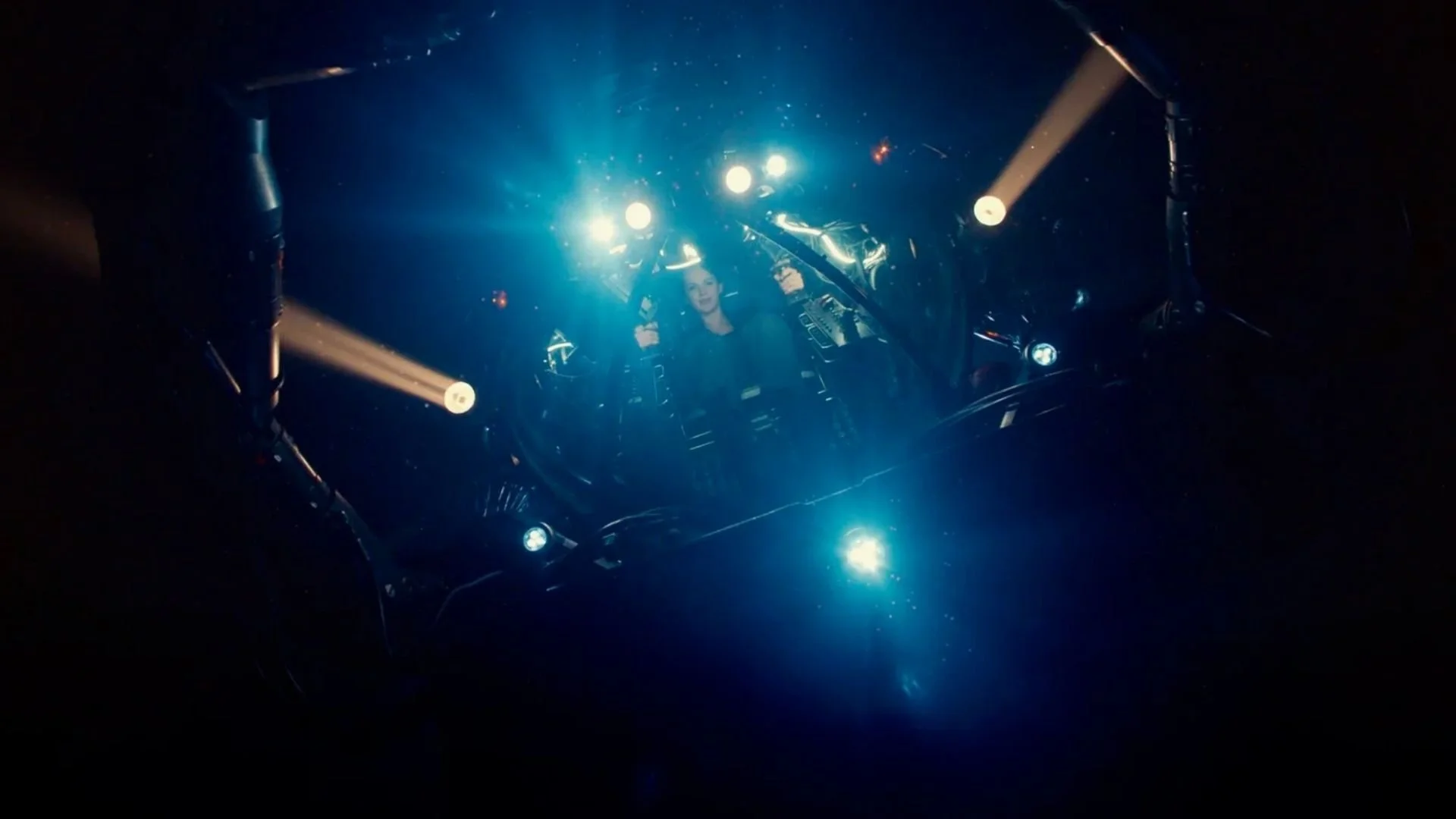 A person inside a cockpit with bright lights and various controls, viewed from above in a dark environment, resembling a spacecraft or aircraft cockpit.