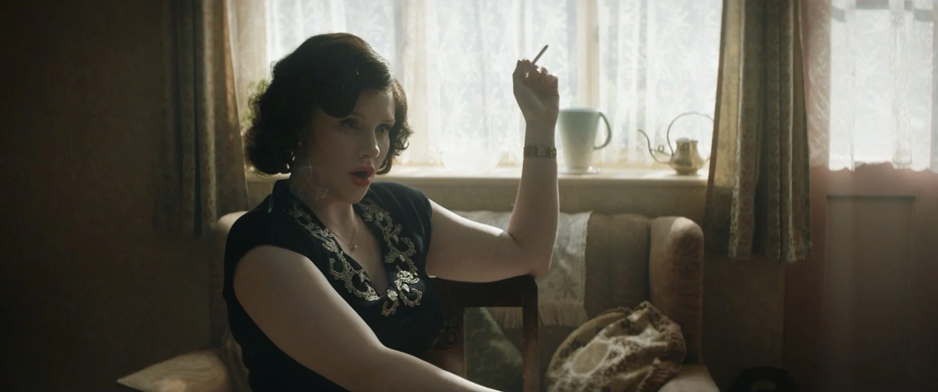 A woman with dark hair in vintage style, wearing a black dress with gold embroidery, sitting on a beige sofa in a room with floral curtains and lace-window coverings, holding an object in her right hand and looking surprised.