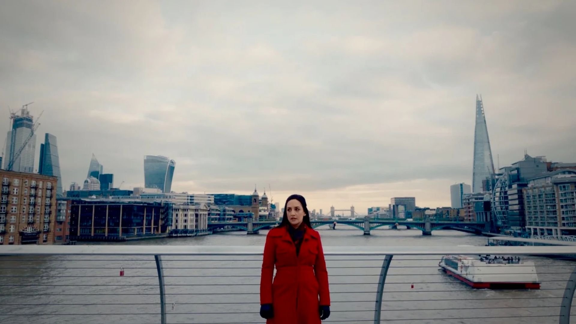 A woman in a red coat standing on a bridge over the Thames River with the London skyline, including buildings like The Shard, visible in the background on a cloudy day.