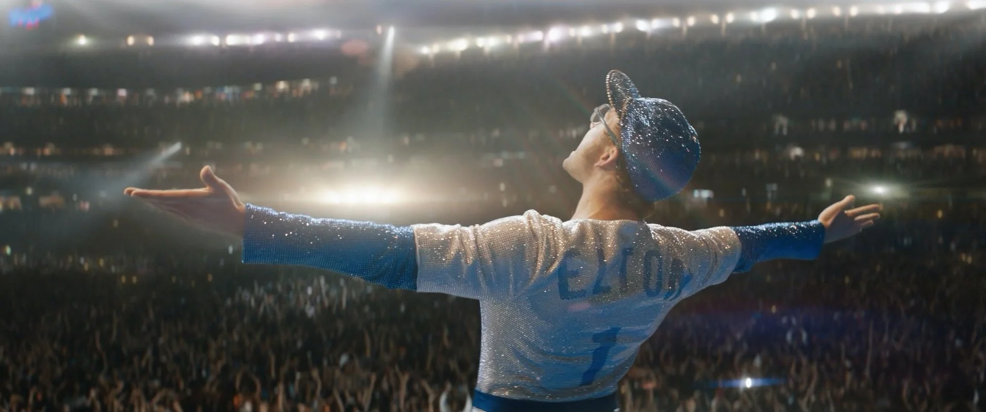Performer on stage with arms outstretched, wearing a shiny silver jacket and a sparkling blue hat, facing a large crowd in an arena.