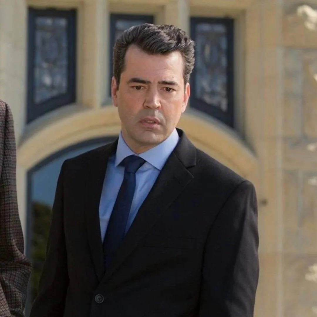 A man in a black suit with a blue shirt and tie standing in front of a building with arched windows, looking serious.