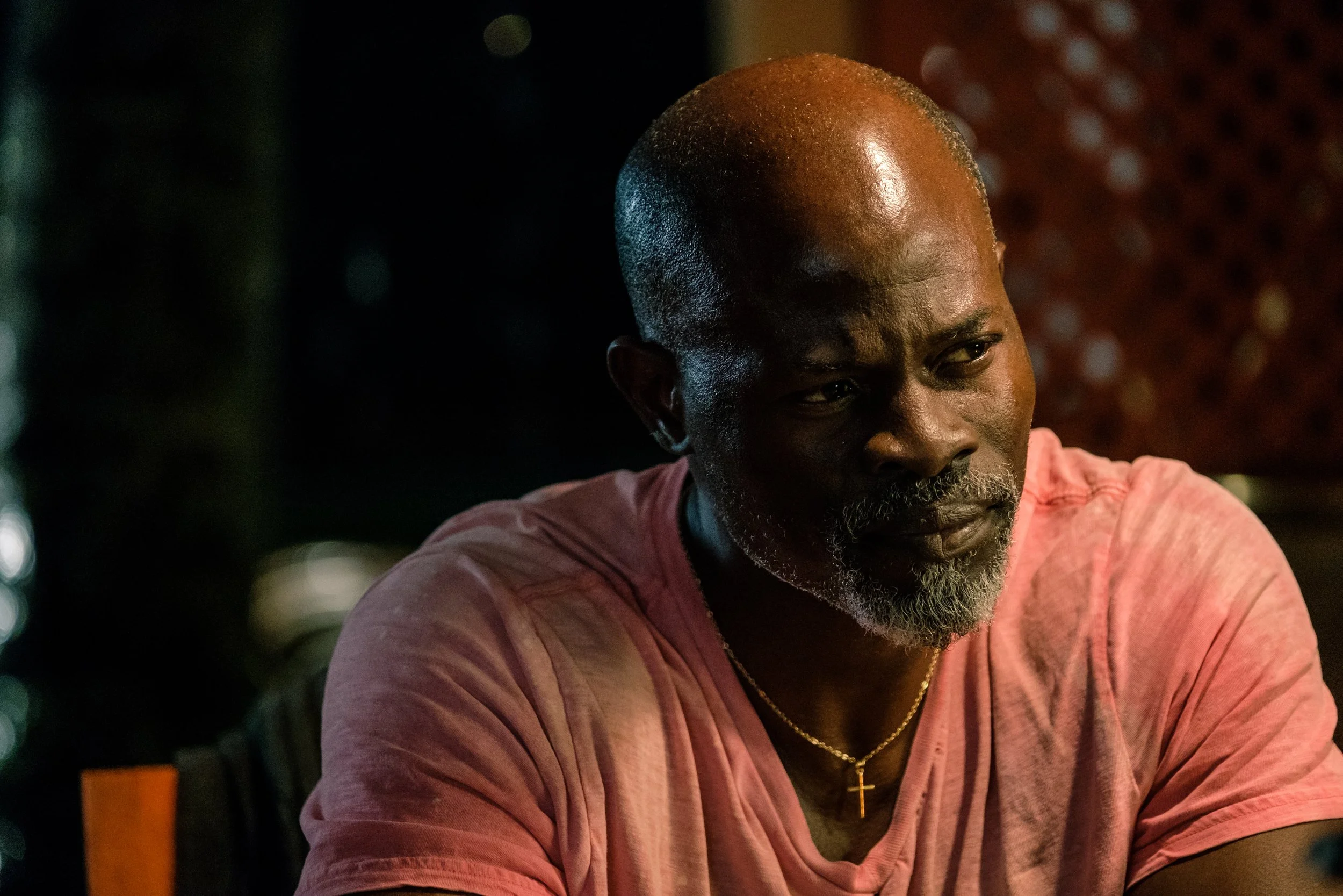 A man with a shaved head and a beard, wearing a pink shirt and a gold chain with a cross, looking slightly to the side, in a dimly lit setting.