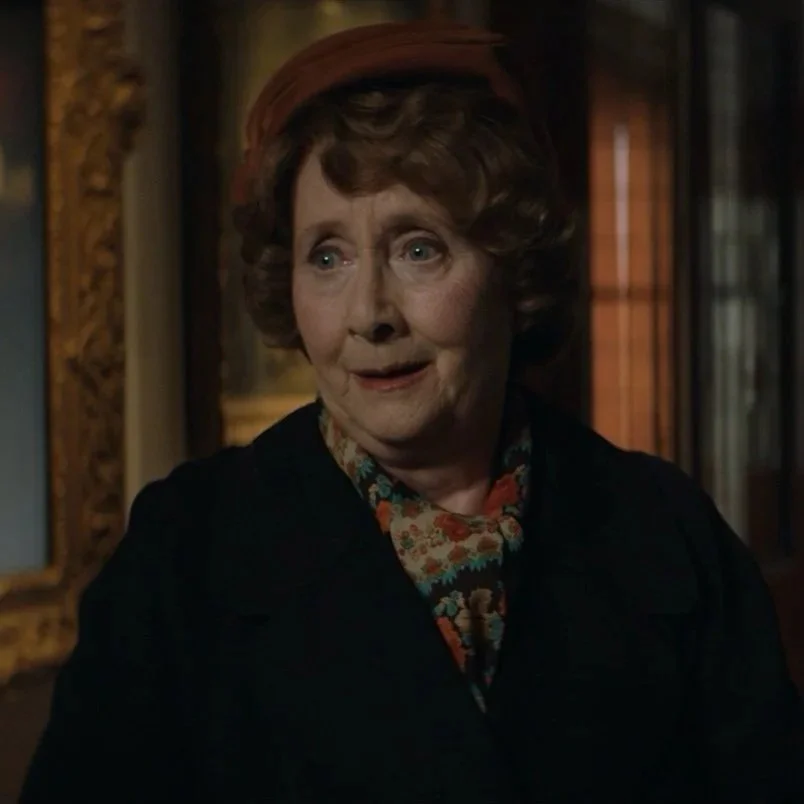 An elderly woman with curly hair, wearing a red hat, floral scarf, and dark jacket, looks surprised or intrigued in an indoor setting with warm lighting and wooden decor.