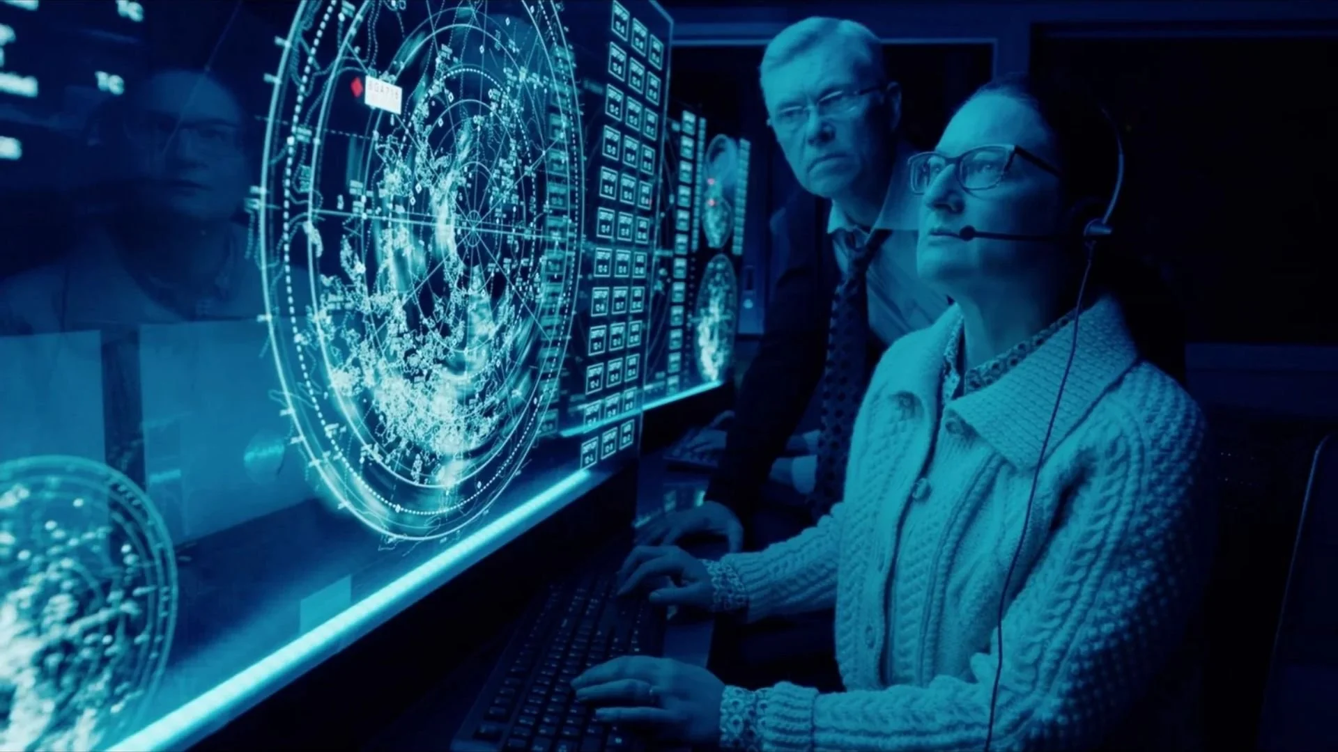 Two people working at a computer with a holographic globe and digital data display, in a high-tech control room or command center.