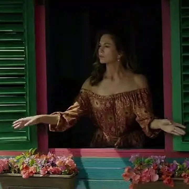 A woman with shoulder-length hair, wearing an off-the-shoulder patterned blouse, leans out of a colorful window with green shutters, extending her right hand while surrounded by flower boxes with pink and purple flowers.