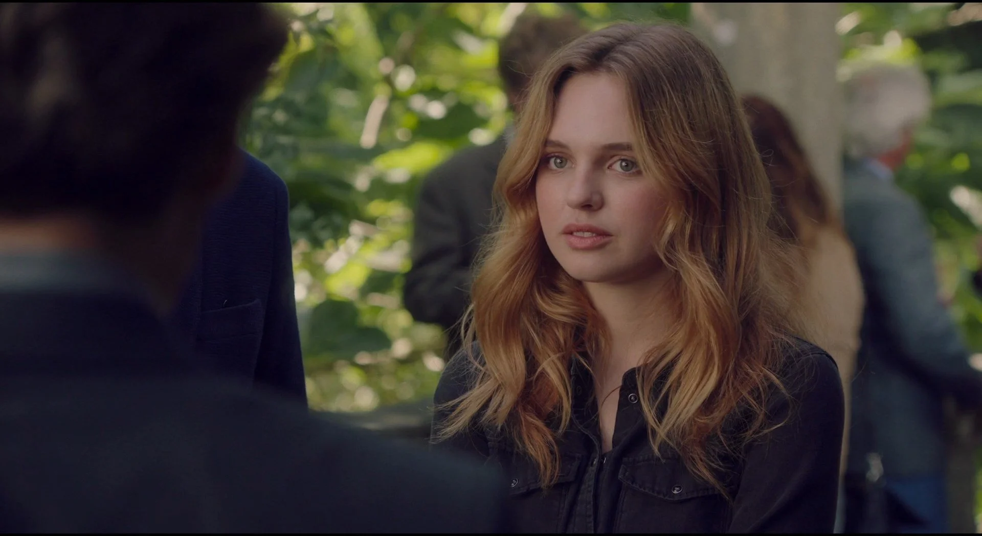 A young woman with wavy red hair and blue eyes, wearing a black jacket, is outdoors surrounded by greenery and is looking at a person out of frame.