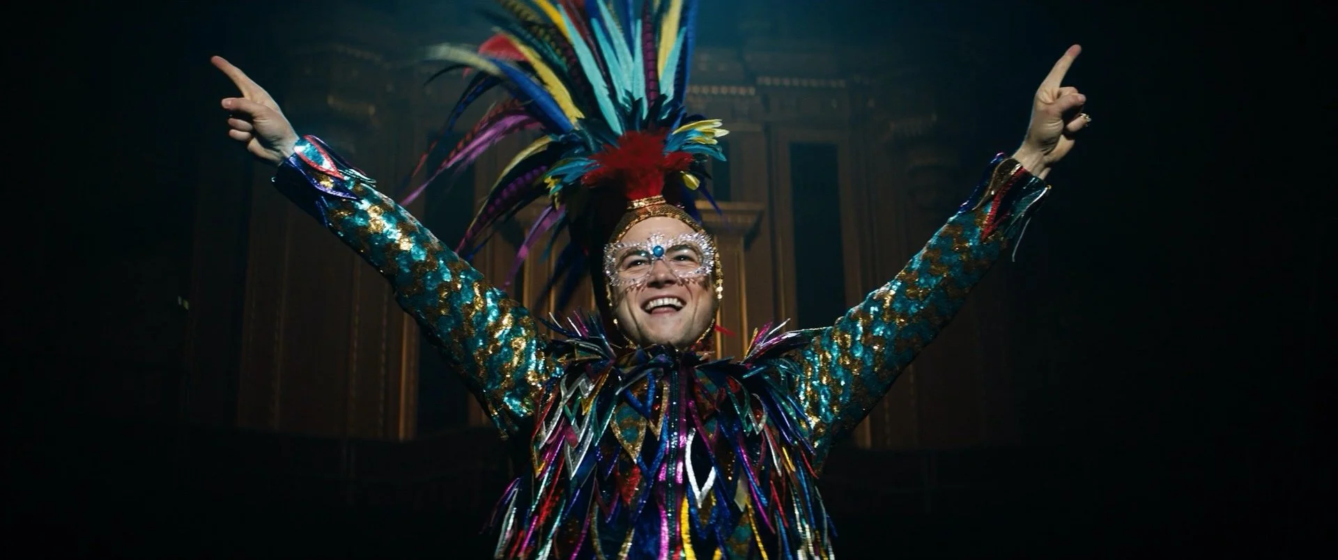 Person wearing a colorful, sequined costume with a feathered headdress and decorative face glasses, smiling with arms raised in a victory pose on a stage.