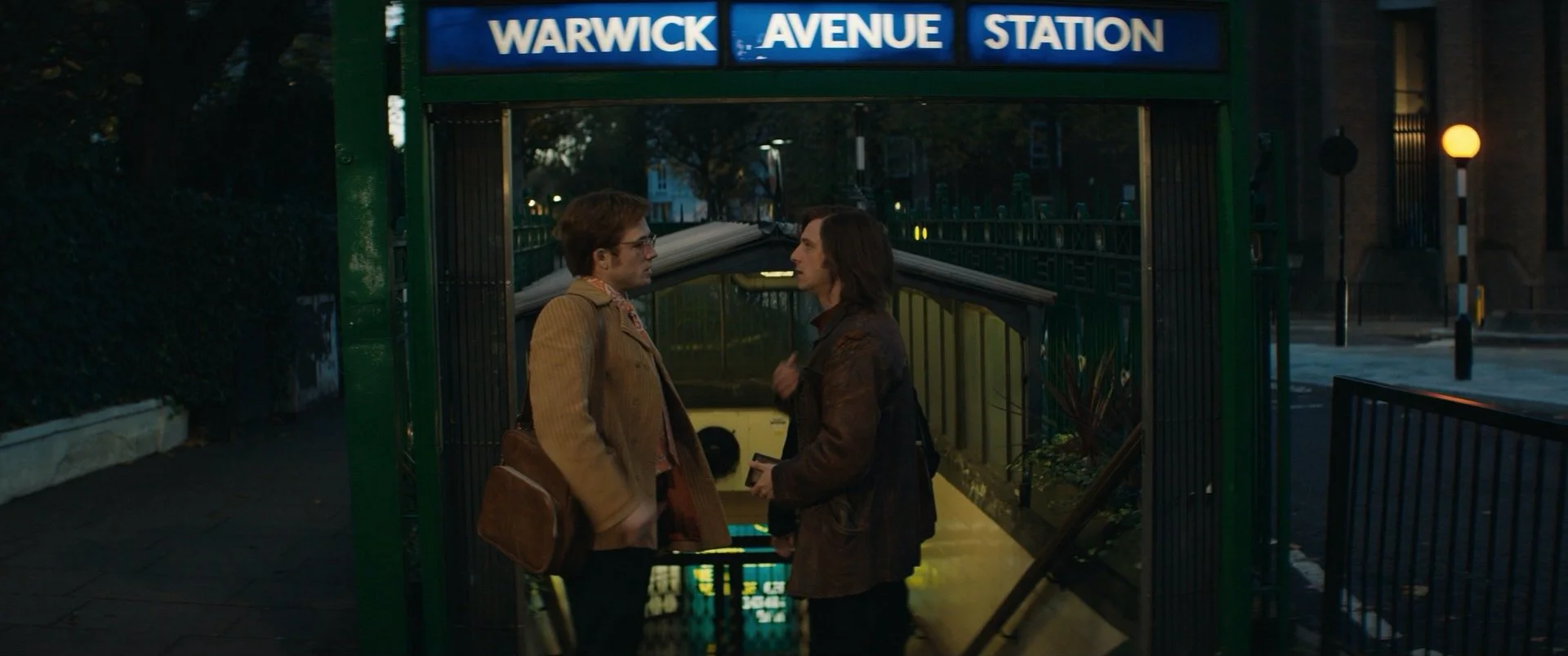 Two men face each other and speak inside a subway station entrance at Warwick Avenue, with the station sign visible at the top of the image.