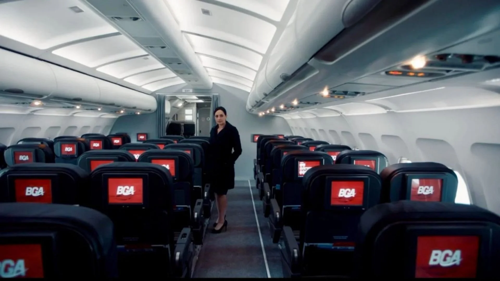 Interior of an airplane cabin with rows of black seats featuring red screens displaying BGA, and a woman in business attire standing in the aisle.