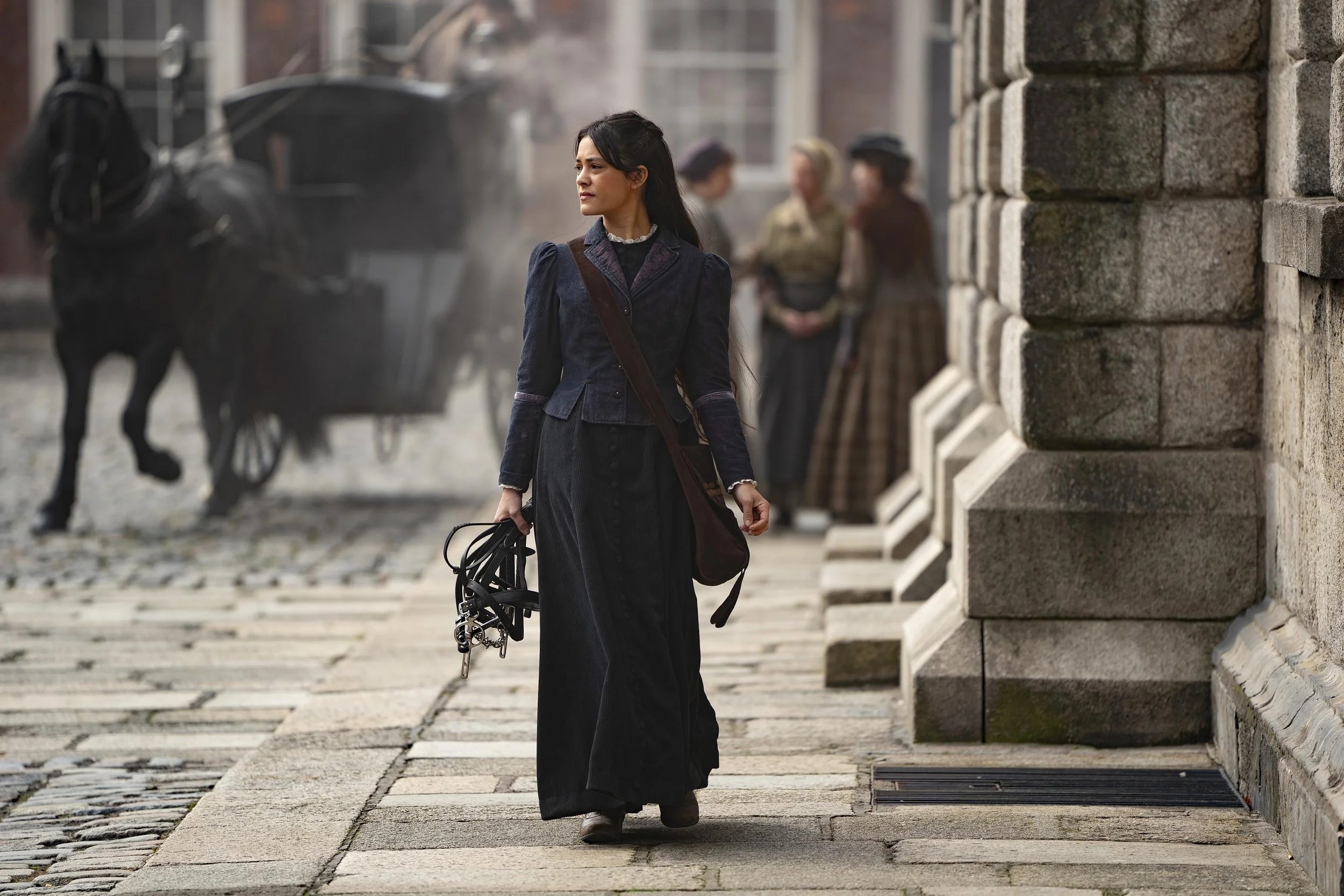 A woman wearing a black Victorian dress holding a bicycle, walking near a stone wall with historical women in period clothing in the background.