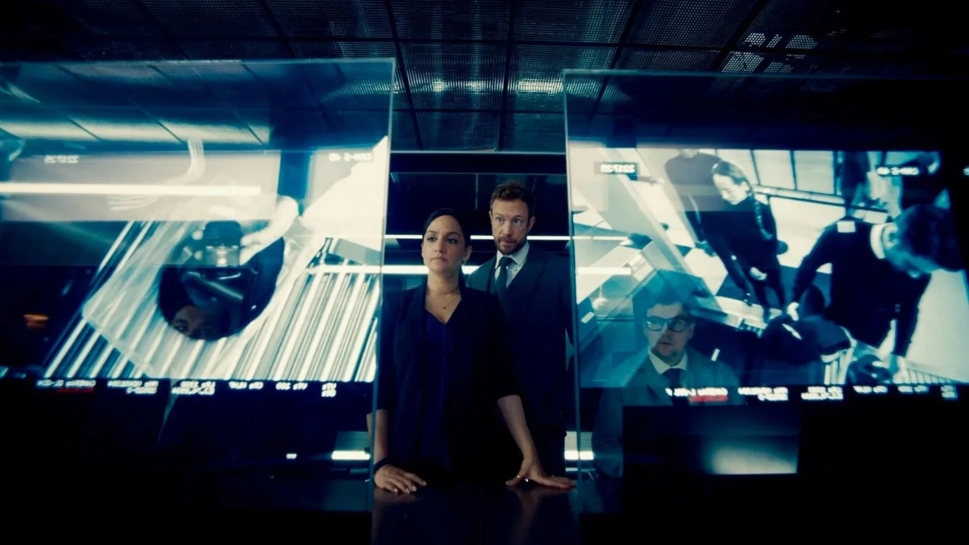 Three people stand in front of a large screen displaying futuristic visuals and data at what appears to be a control room or command center.