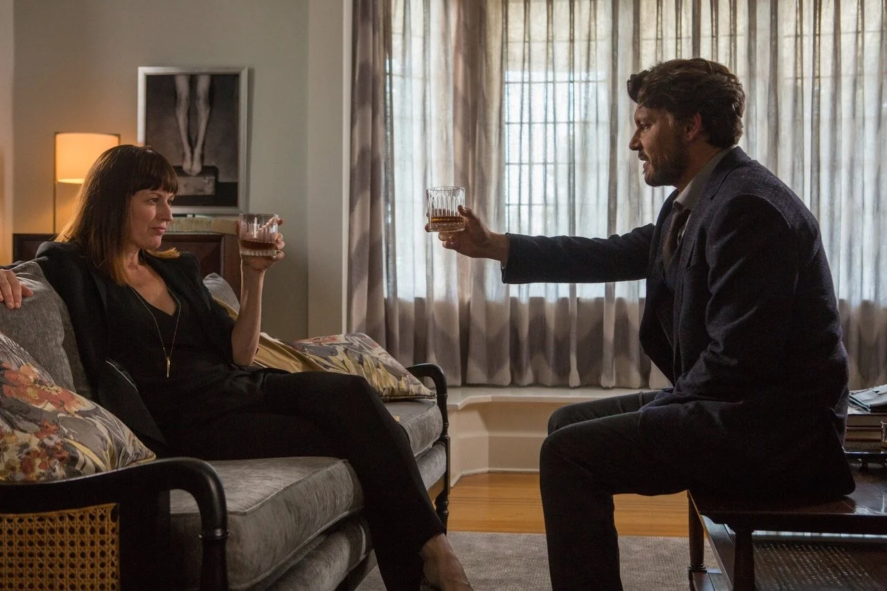 A man and woman having drinks in a living room with a couch, window with curtains, and a lamp in the background.