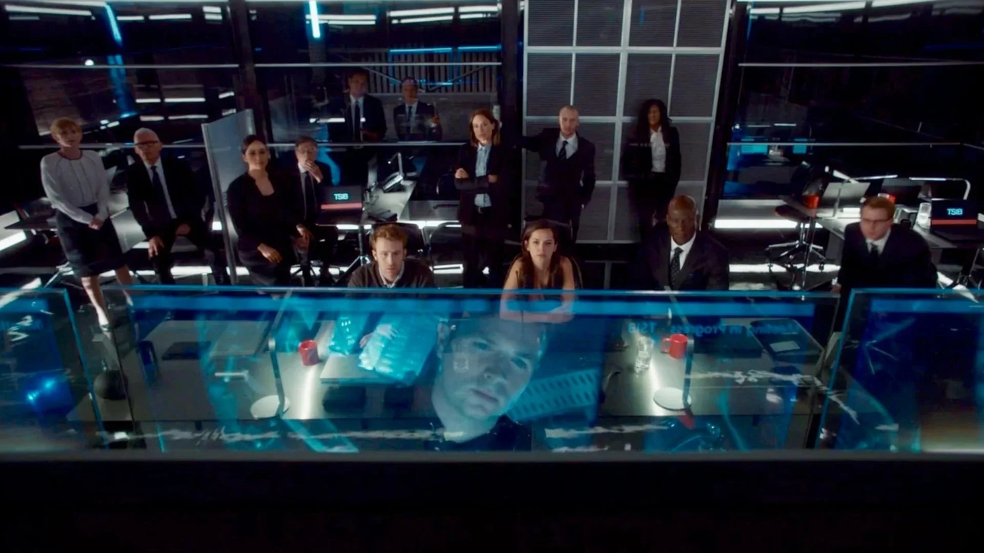 Group of people in a high-tech control room looking at a large transparent screen with a face projected on it.