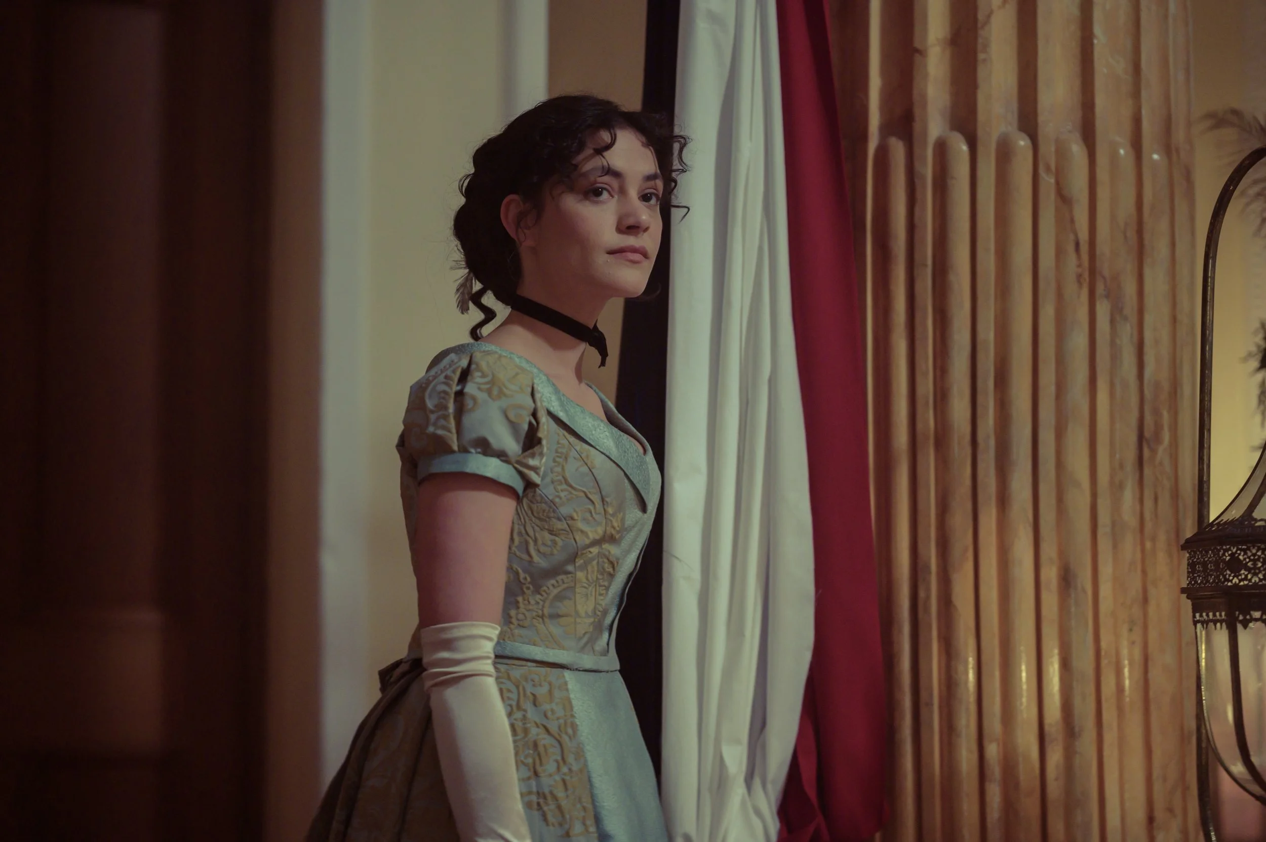 A woman dressed in historical attire looking out from behind curtains with a wooden wall and mirror in the background.