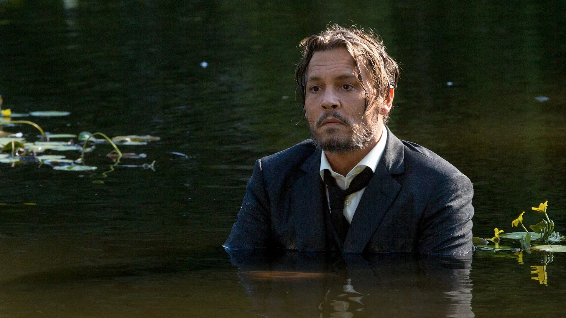 A man with wet hair and a beard wearing a black suit and white shirt, sitting in a body of dark water with yellow flowers, with a serious expression and looking to the right.