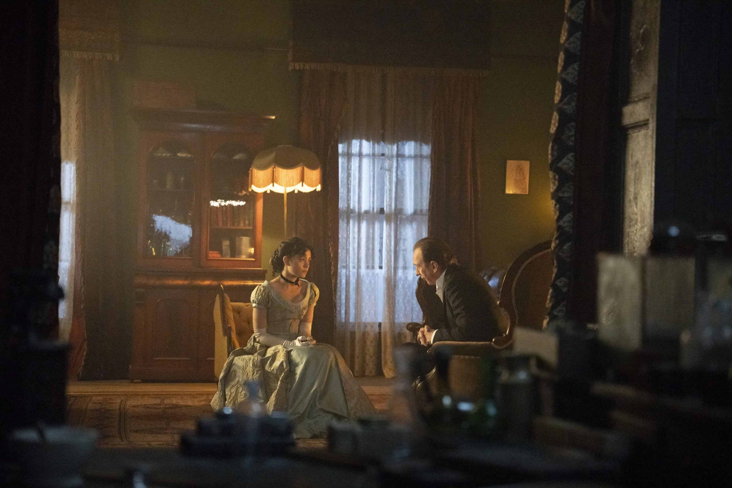 A young woman dressed in a vintage-style gown and gloves sitting on a chair, talking to an older man in a suit inside a dimly lit, vintage-styled room with curtains, a wooden cabinet, and a lamp.