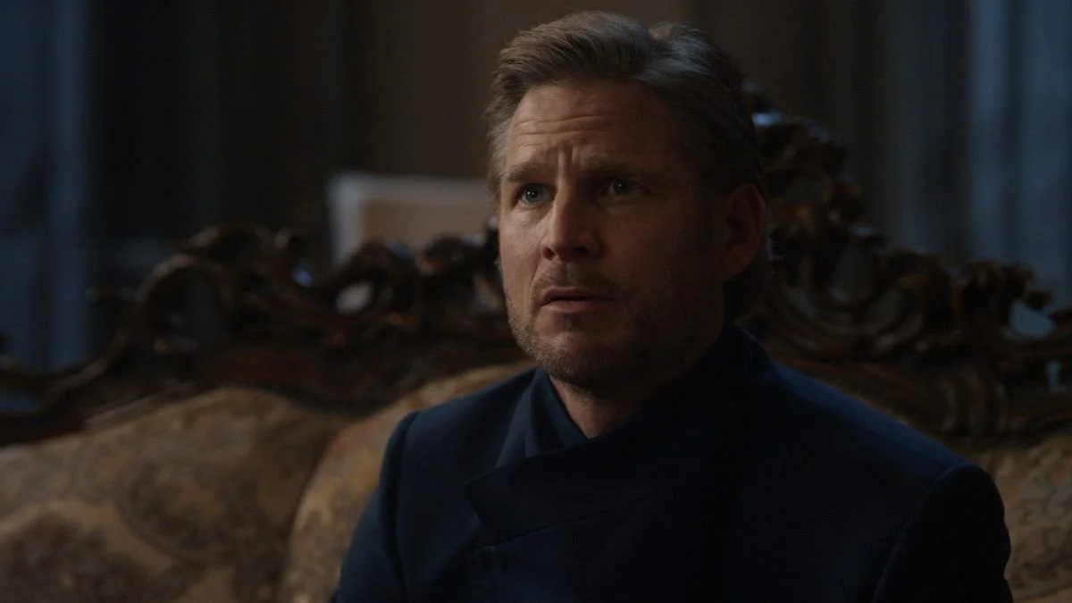 A middle-aged man with brown hair and a stubble beard, wearing a dark suit, sitting in a dimly lit room with ornate wooden furniture in the background.