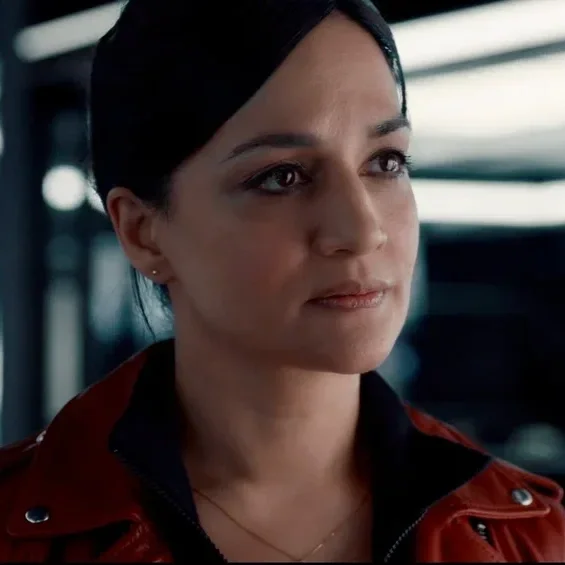 Close-up of a woman with dark hair, wearing a red leather jacket, looking thoughtful indoors.