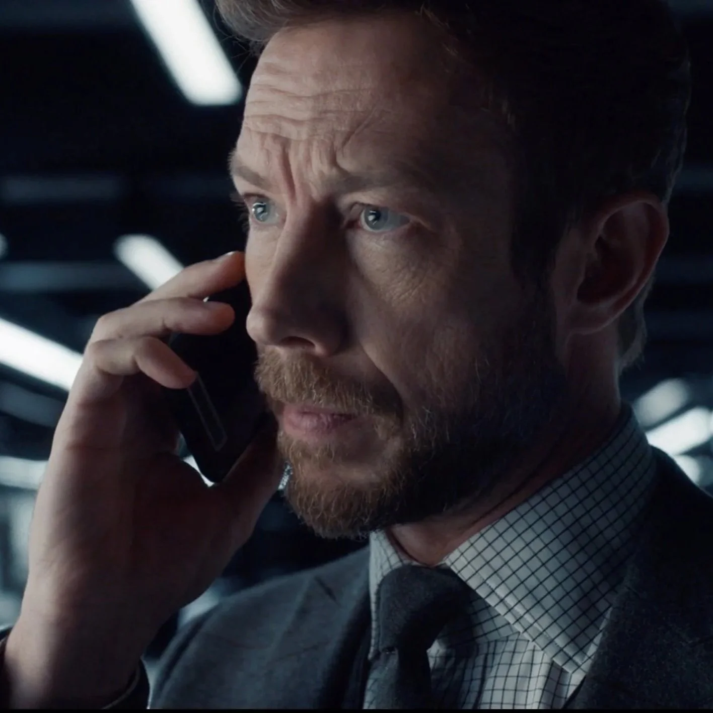 A middle-aged man with blue eyes, a reddish beard, and brown hair, wearing a checked shirt and black tie, talks on a cell phone in a modern indoor setting with bright lighting.