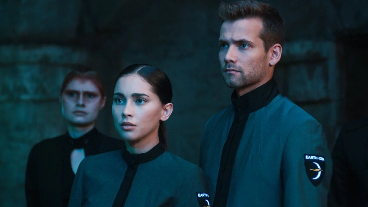 Three individuals in dark uniforms with NASA-like patches, standing in a dimly lit, stone-walled environment, looking serious and focused.