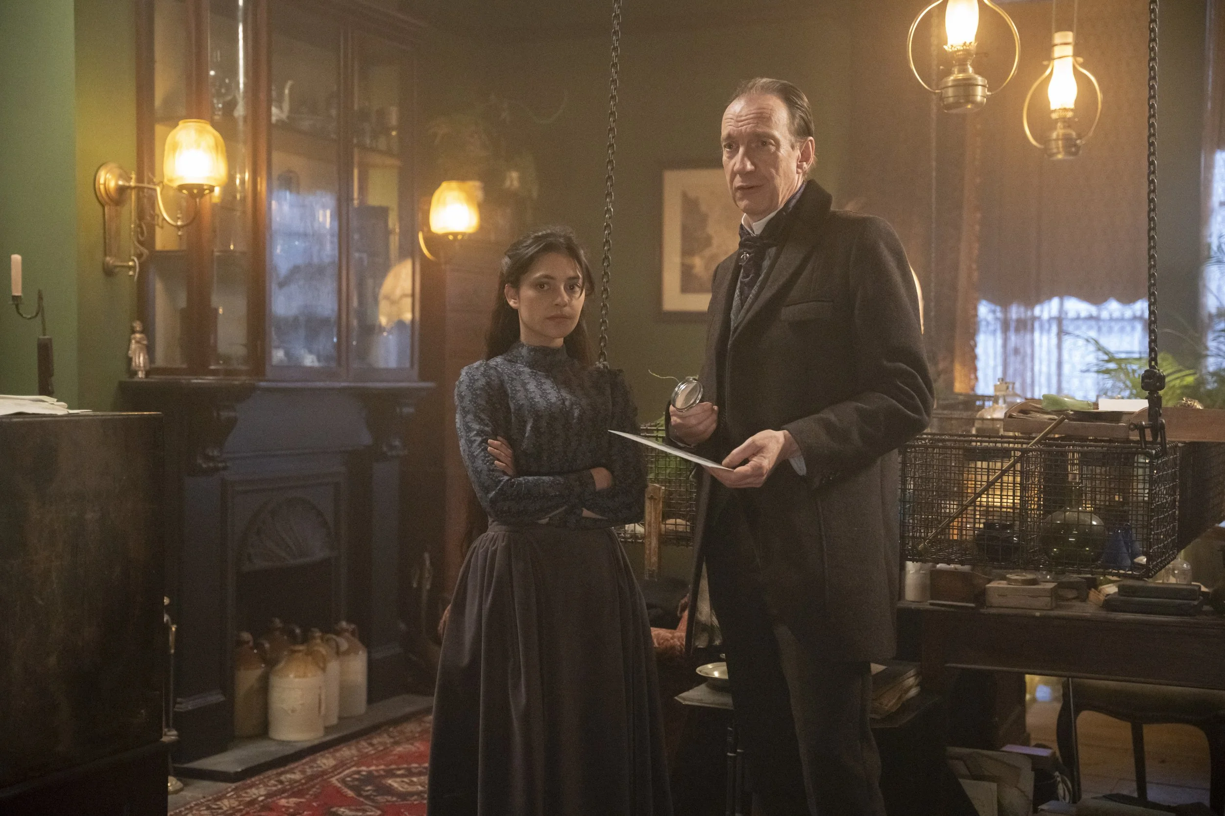 A man and a woman standing in a vintage-style room with warm lighting, dark wood furniture, and a patterned rug. The woman is crossing her arms and looking serious, while the man is holding a key and looking to the side.