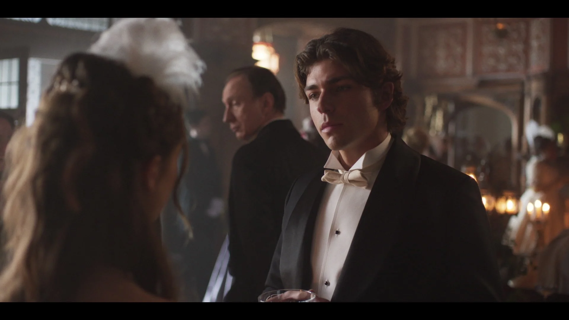A scene set in a fancy, ornate room with a young man dressed in formal tuxedo, holding a drink, looking at a woman in a dress with a feathered hat, amidst other well-dressed guests.