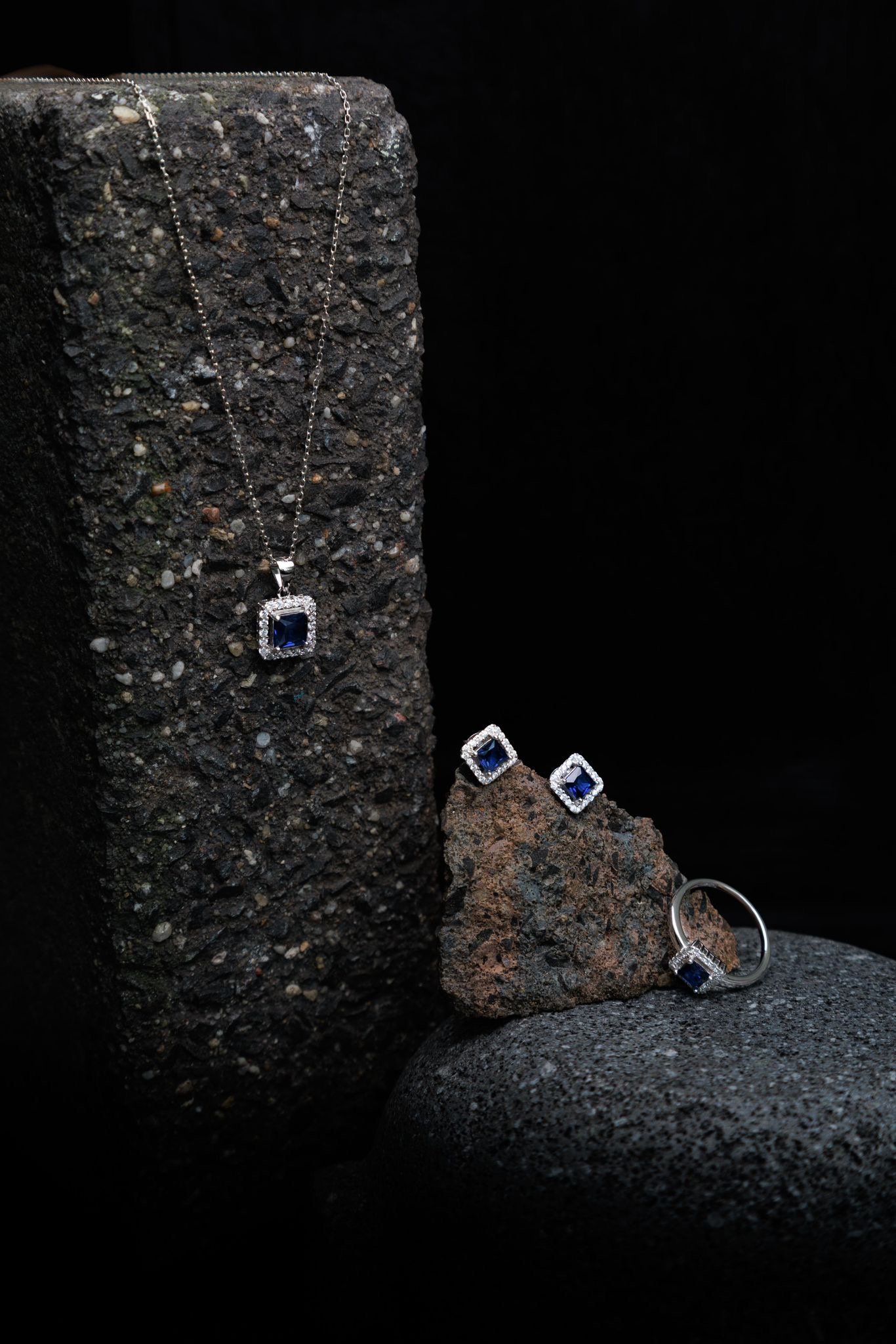 Set of jewelry including a necklace, earrings, and a ring, all featuring square-shaped blue gemstones with surrounded by small clear stones, displayed on rough, dark textured rocks against a black background.