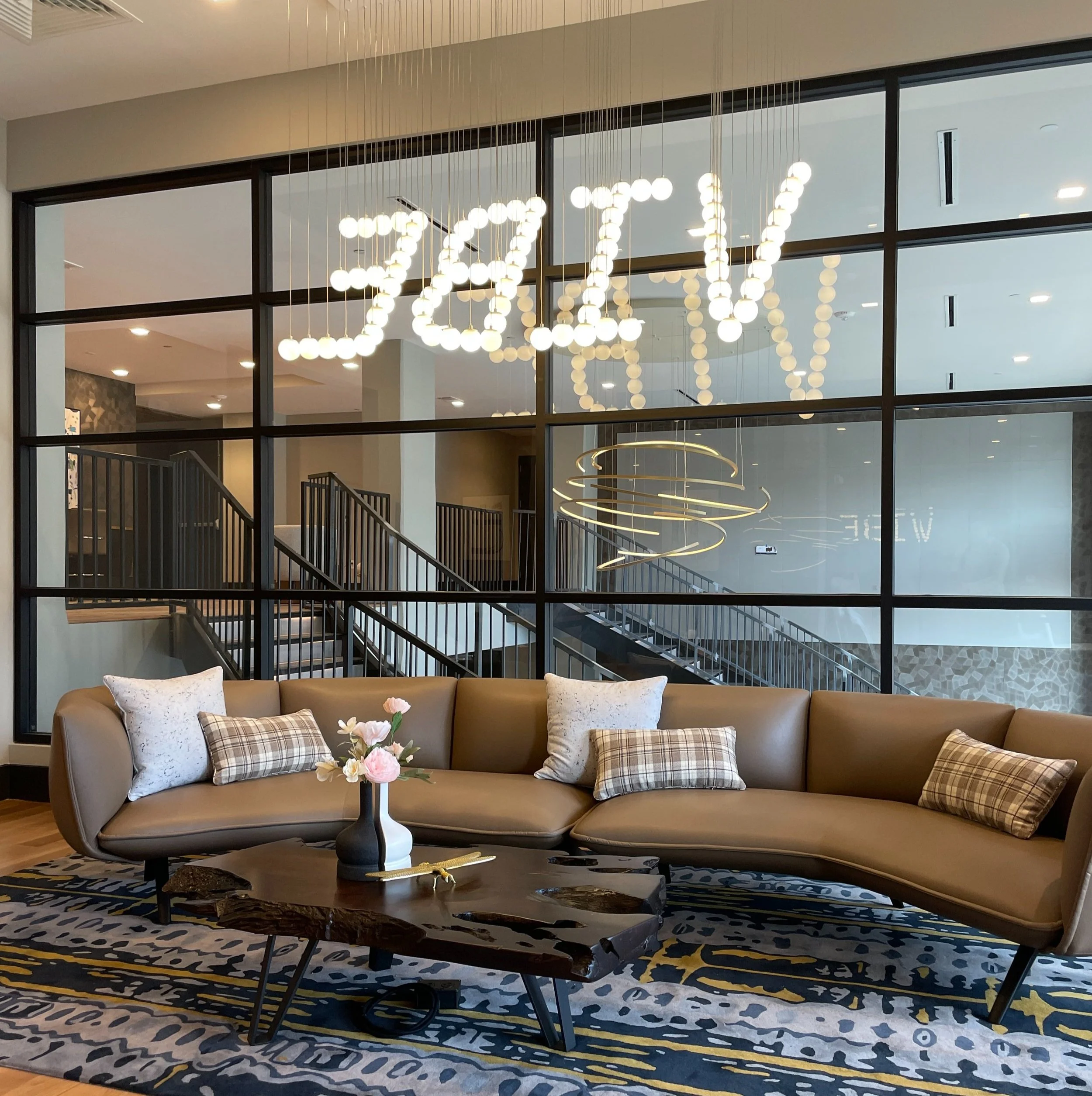 A modern lobby with a curved beige sofa adorned with plaid and white pillows. In front, a dark wooden coffee table holds a vase with pink and white flowers. Behind the sofa, a large glass wall features decorative light fixtures. The space has a stair