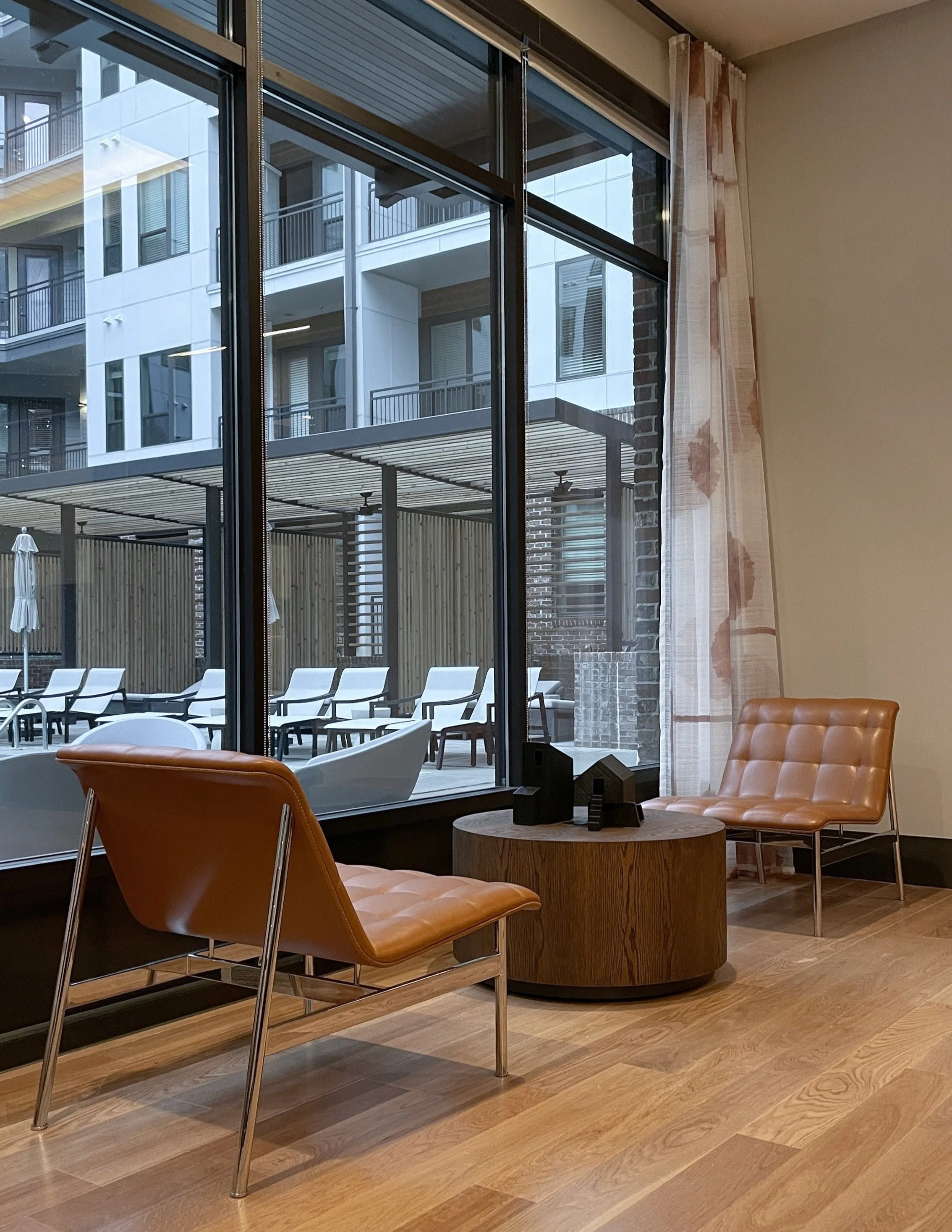 Interior view of a lounge area with tan leather chairs, a round wooden table with decorative objects, beige carpeting, and a large window revealing a poolside with lounge chairs and a modern building outside.