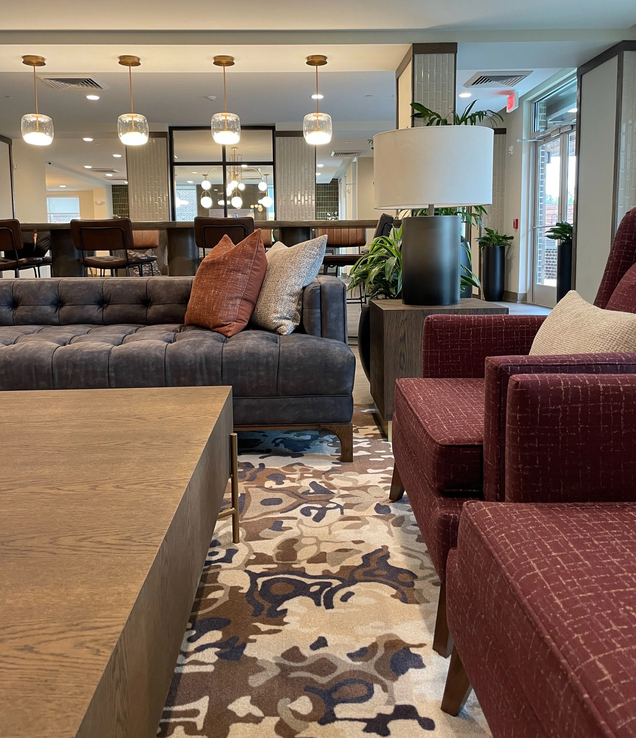 Interior view of a modern lounge area in an apartment, featuring a dark tufted sofa with pillows, red patterned chairs, a wooden table, decorative plants, and overhead lighting fixtures.