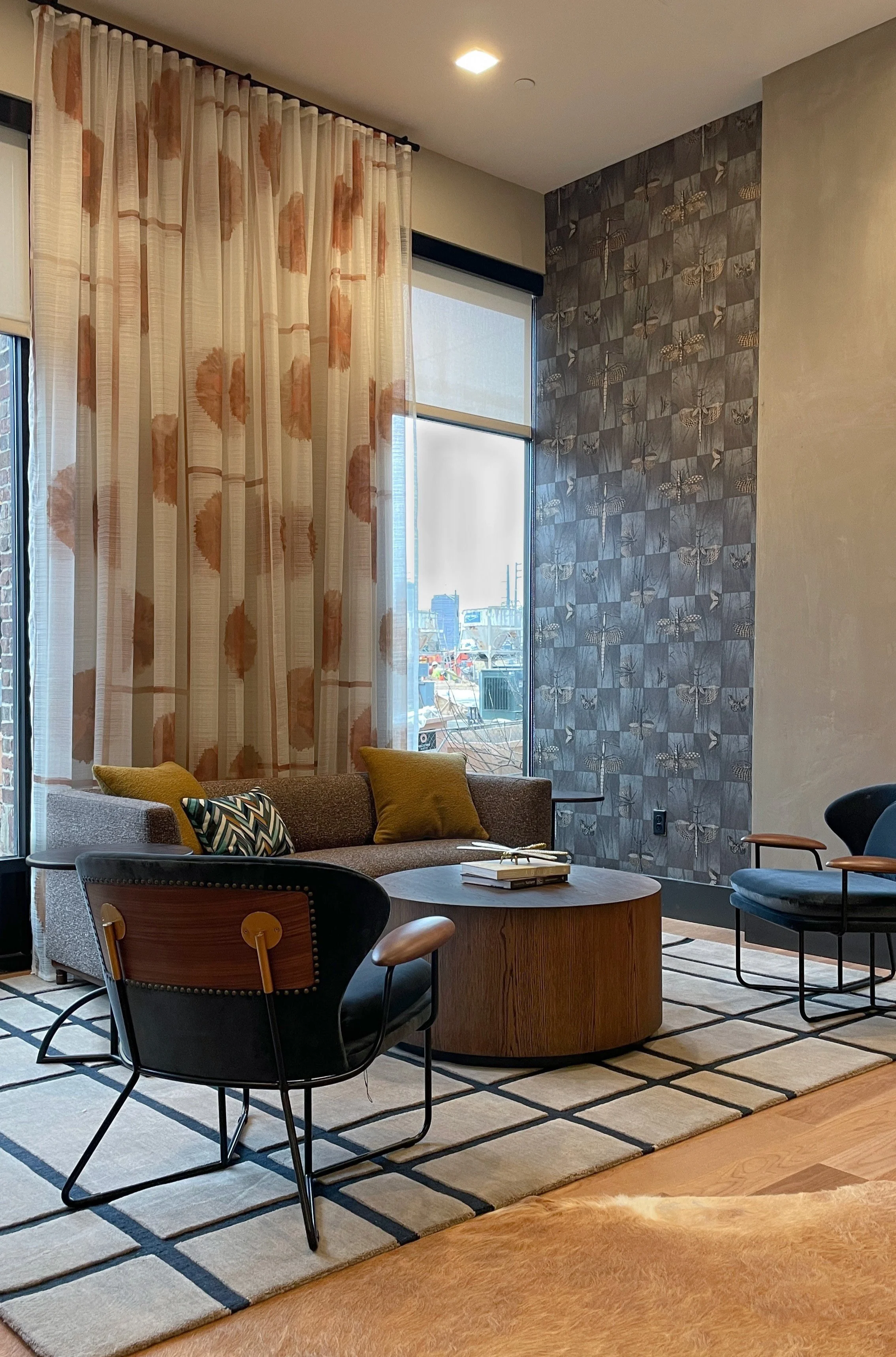 A modern lounge area with a grey sofa, colored cushions, a round wooden coffee table, black armchairs, large windows with patterned curtains, a patterned wallpaper wall, and a geometric patterned rug.