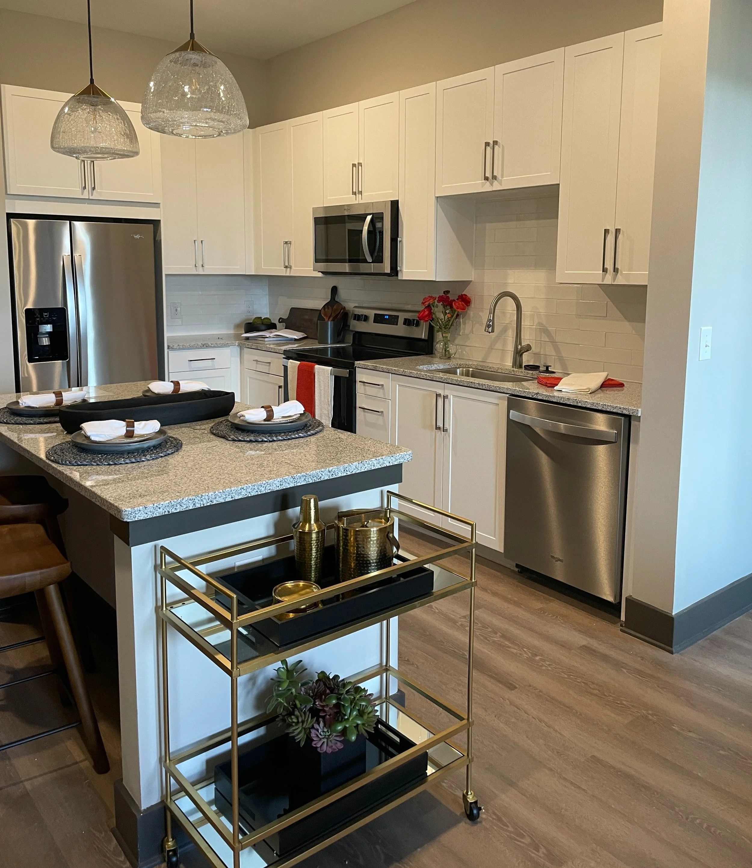 Modern kitchen with white cabinets, stainless steel appliances, granite countertops, and a kitchen island with place settings. There is a gold and black serving cart with decorative items and a potted plant. Pendant lights hang above the island.