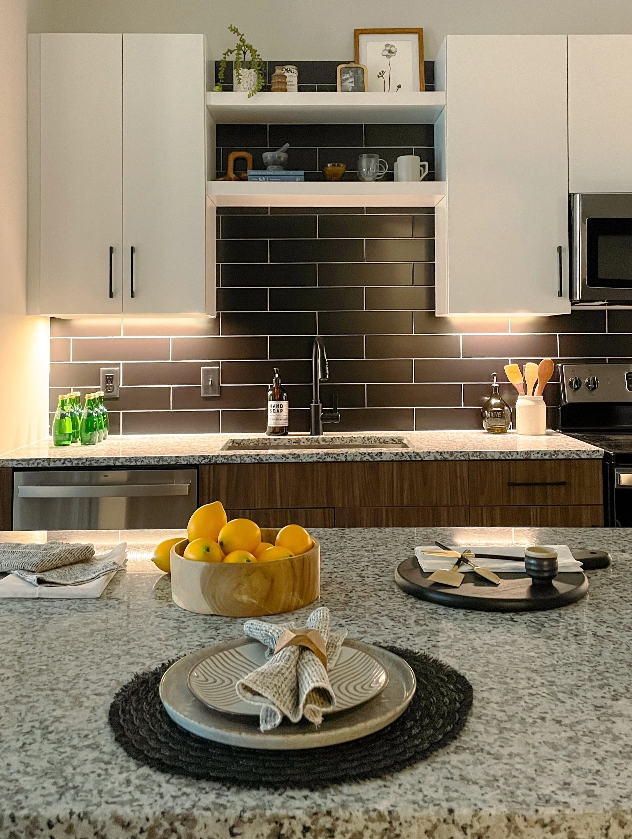 Modern kitchen with granite countertops, black tile backsplash, white cabinets, and open shelves decorated with framed artwork and plants.