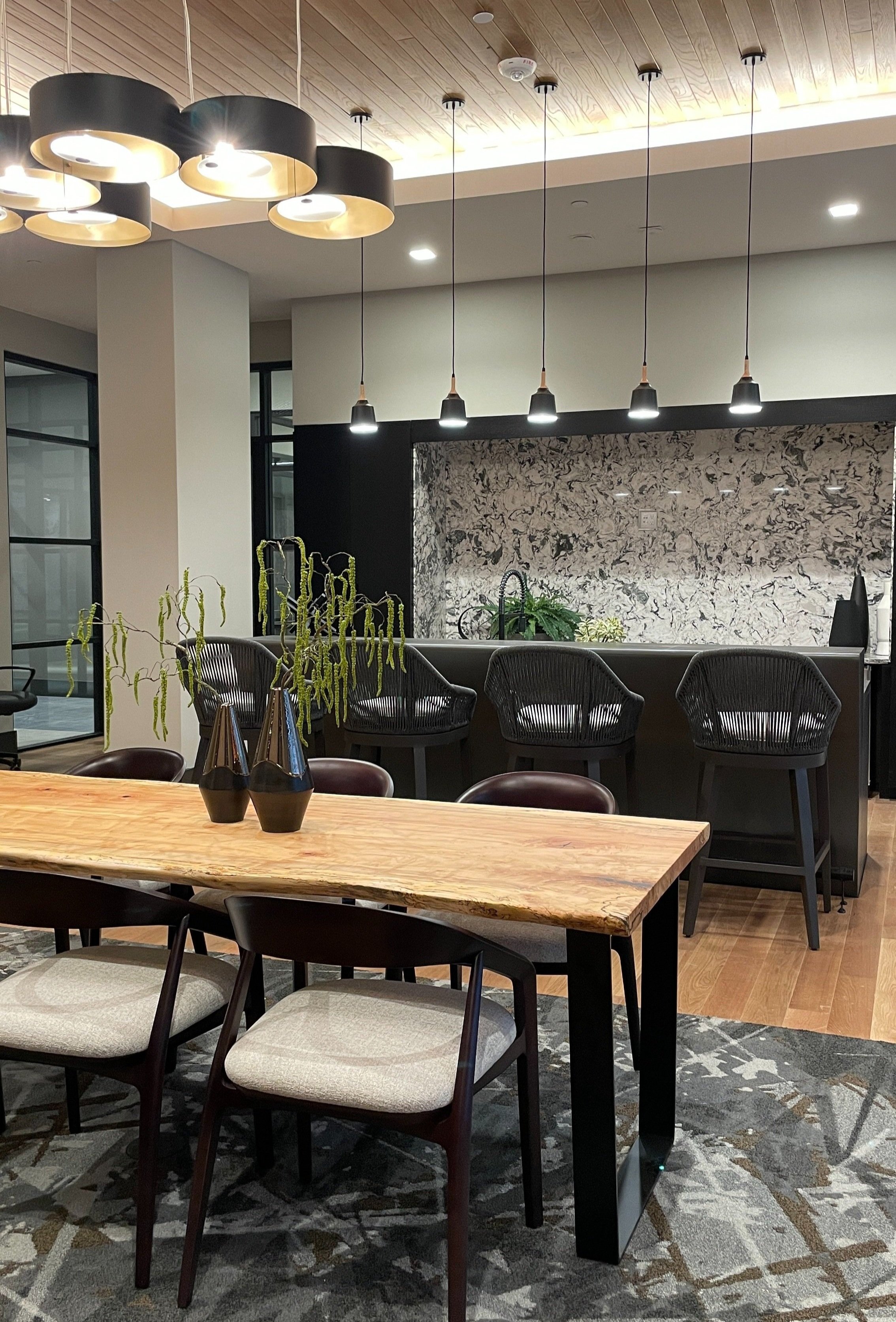 Modern dining area with a wooden table, beige upholstered chairs, black vases with green branches, pendant lights, and a custom pre-cast concrete kitchen island with black chairs in front of a kitchen counter with a marbled backsplash.