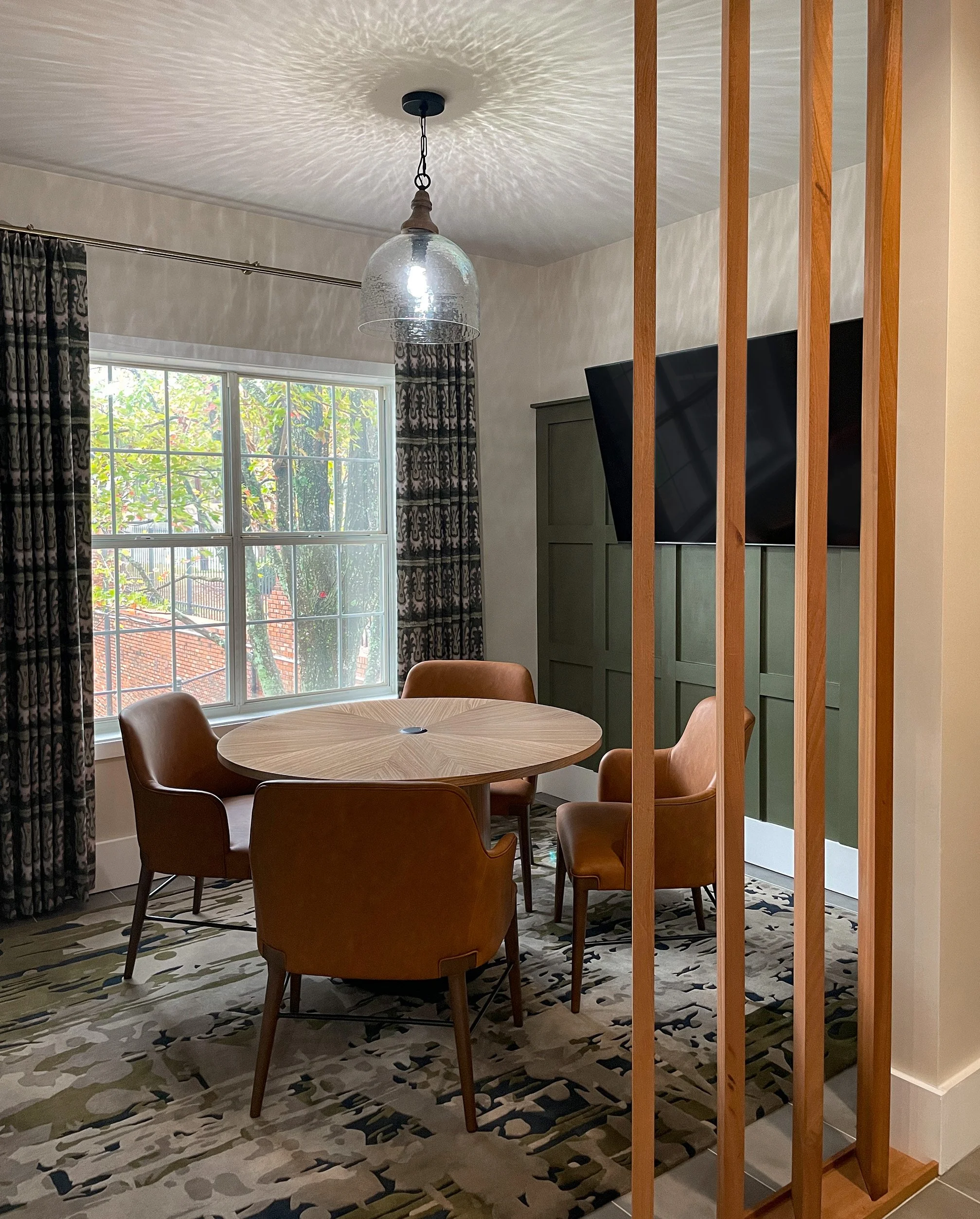 Dining area with a wooden oval table surrounded by five brown leather chairs, a large window with patterned curtains, and a wall-mounted TV, with a wooden room divider to the right.