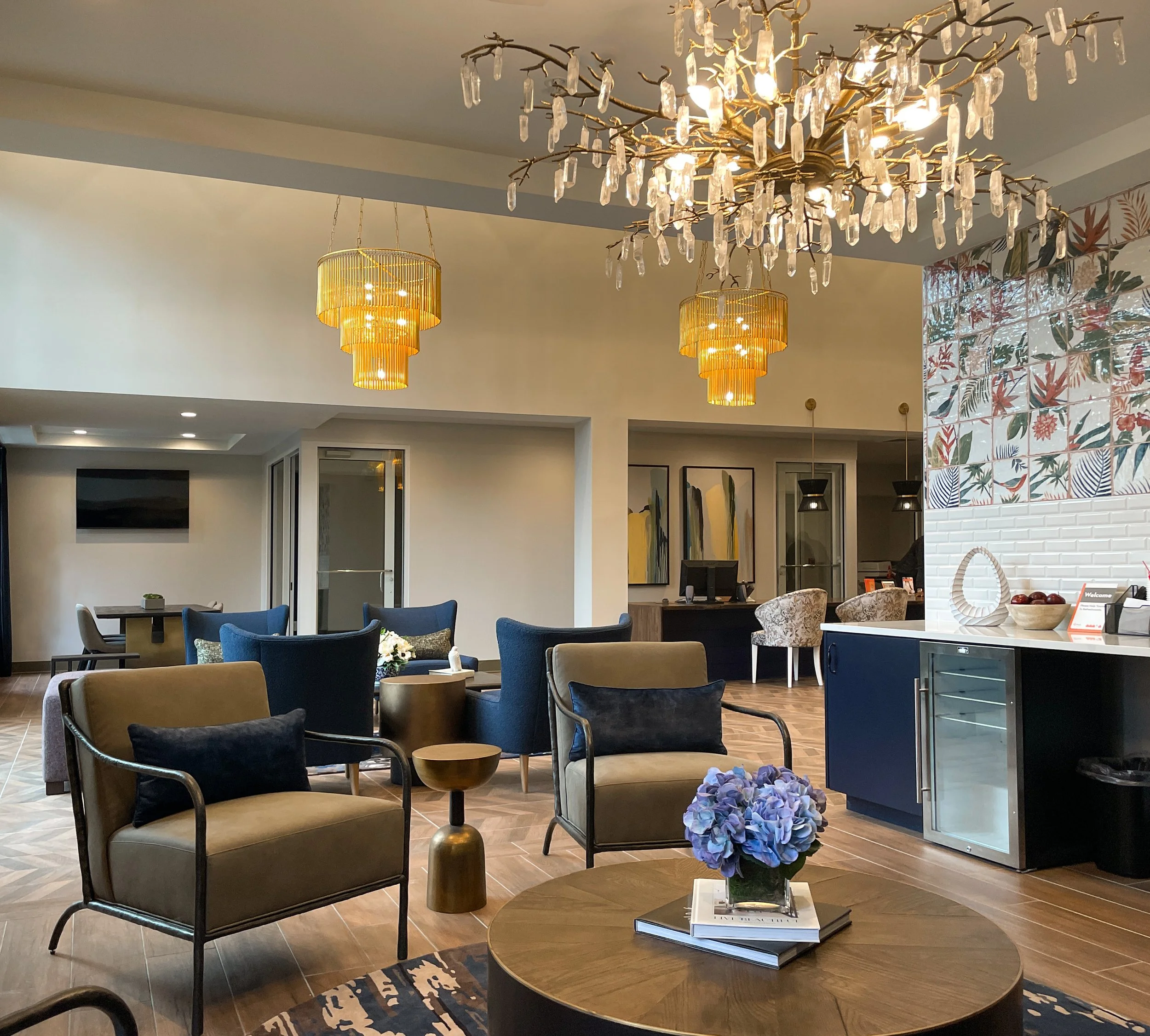 A stylish hotel lobby with modern furniture, decorative lighting, a floral artwork on the wall, and a chandelier with hanging crystals.