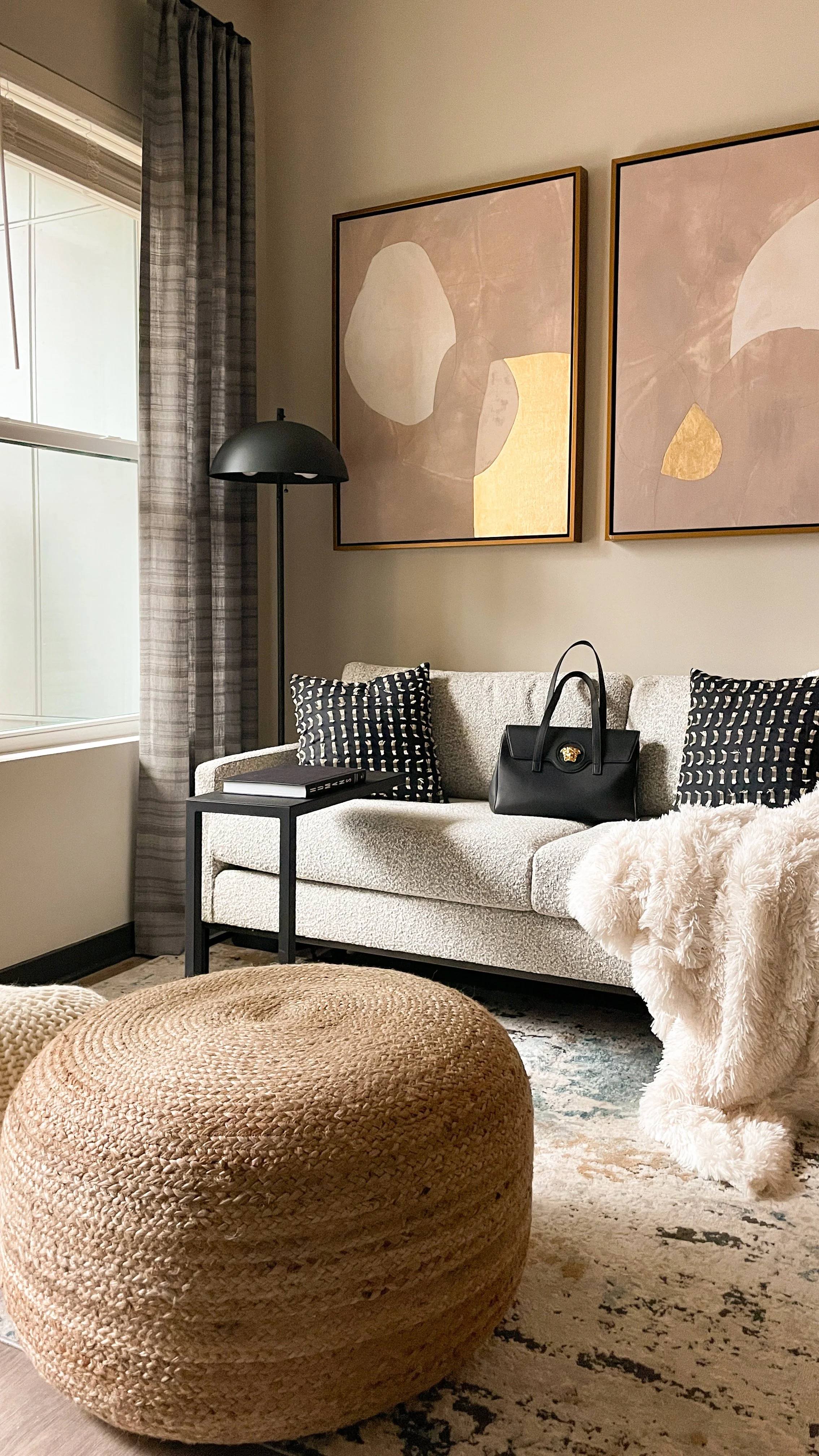 Living room with a beige sofa, black and white patterned pillows, a black handbag, a side table with a closed book, a black floor lamp, two abstract paintings, a window with checkered curtains, a woven pouf, a fluffy throw blanket, and a patterned ru