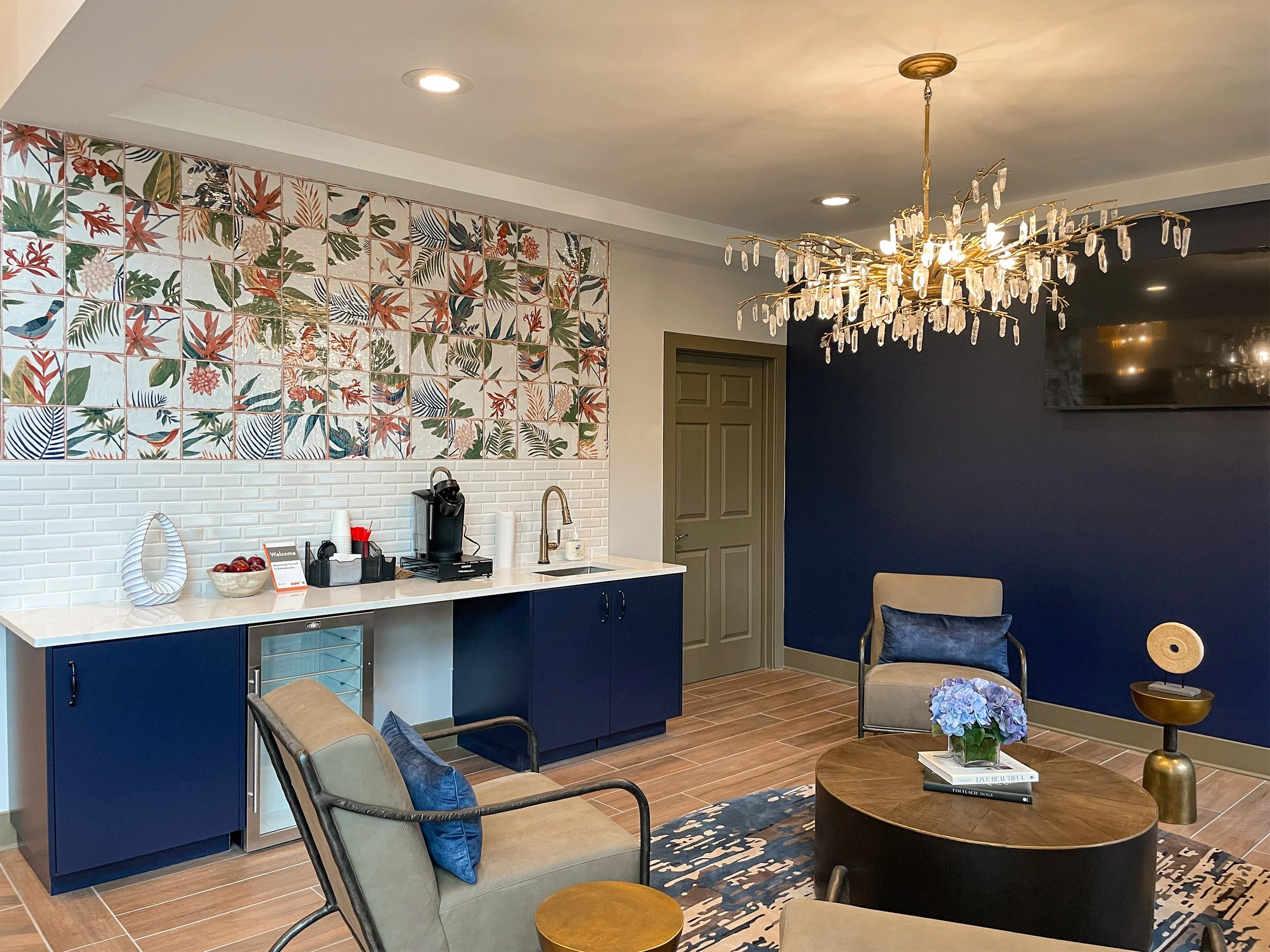 A modern living room with a blue accent wall, a chandelier, and a seating area with beige chairs, a round coffee table with a blue hydrangea bouquet, and decorative items. A colorful botanical tile mural backsplash decorates the kitchenette area.
