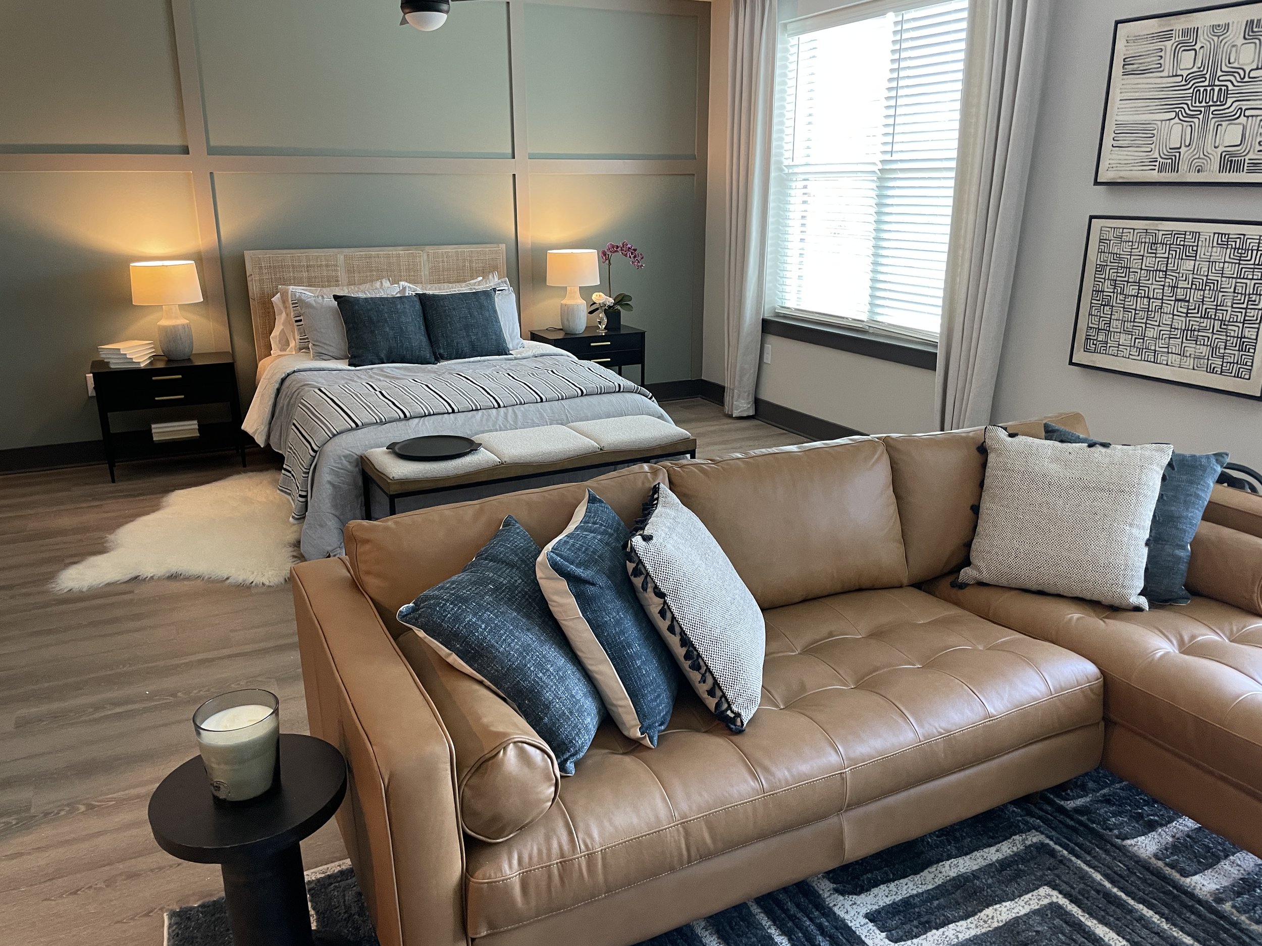 Studio apartment living room with a tan leather sectional sofa with blue and white pillows, a black side table with a candle, a cozy bed in the background with a bench, lamps, and modern artwork on the wall.