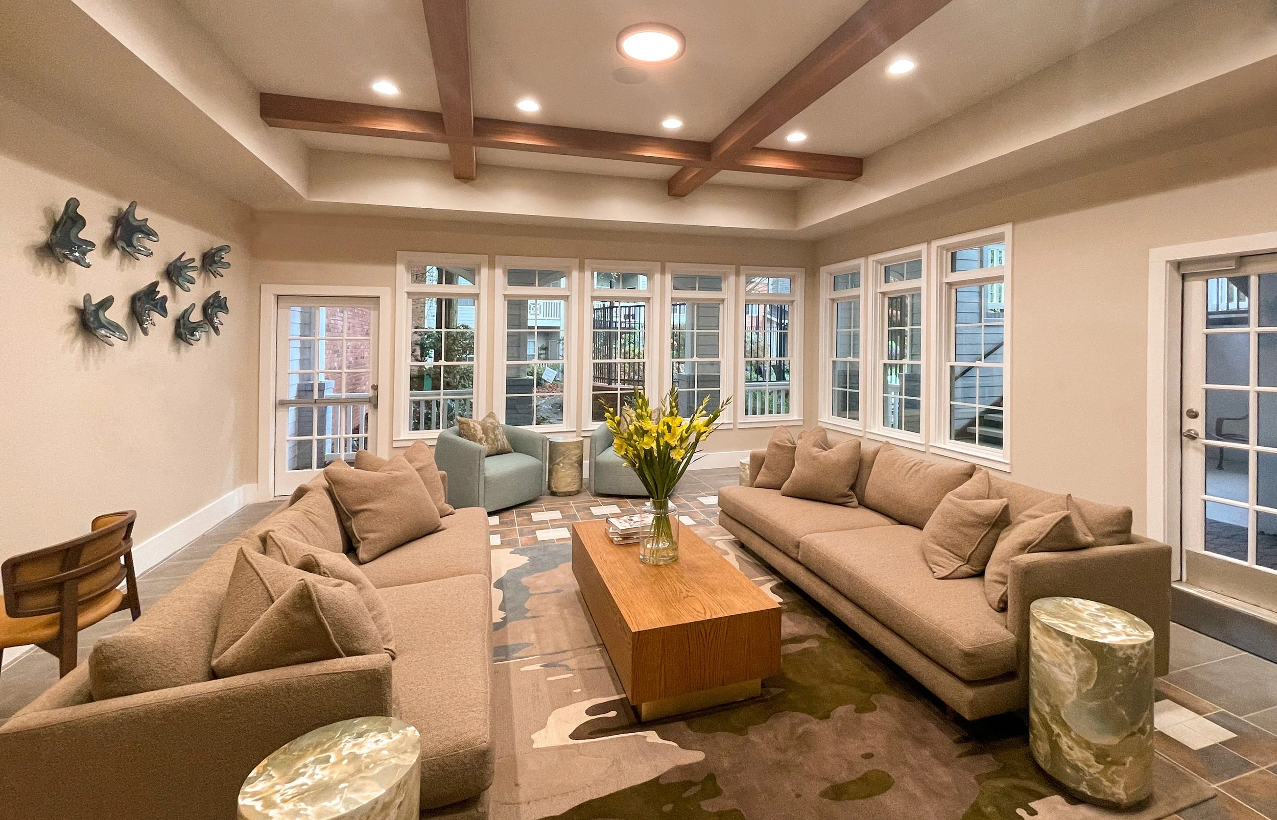 A cozy living room with two beige sofas, a wooden coffee table with a flower vase, several throw pillows, two green armchairs near a large bay window, and decorative wall art of ceramic fish. The room has a tray ceiling with wooden beams, multiple re