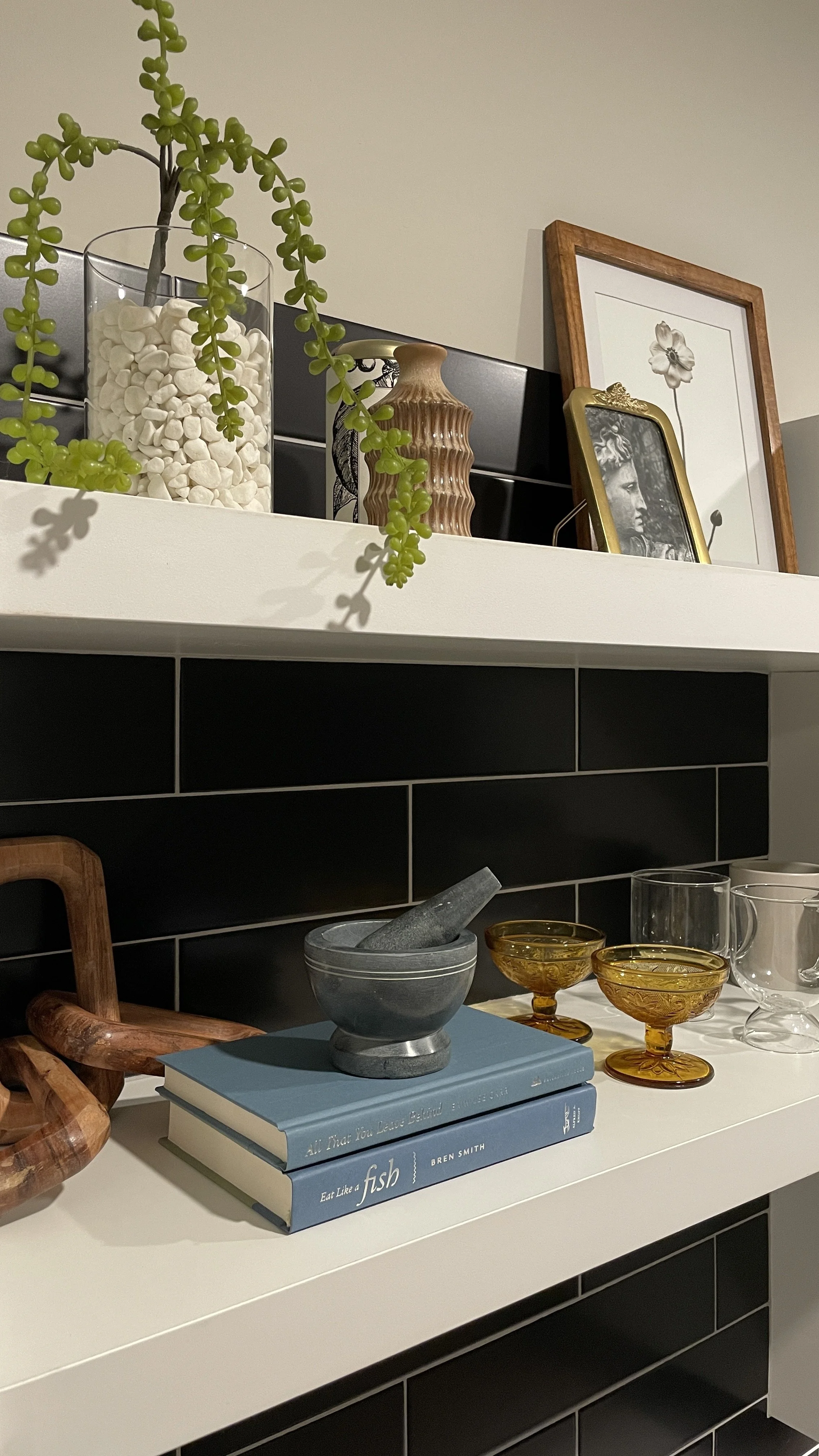 Decorative shelf with plant, framed pictures, books, glassware, and ceramic mortar and pestle.