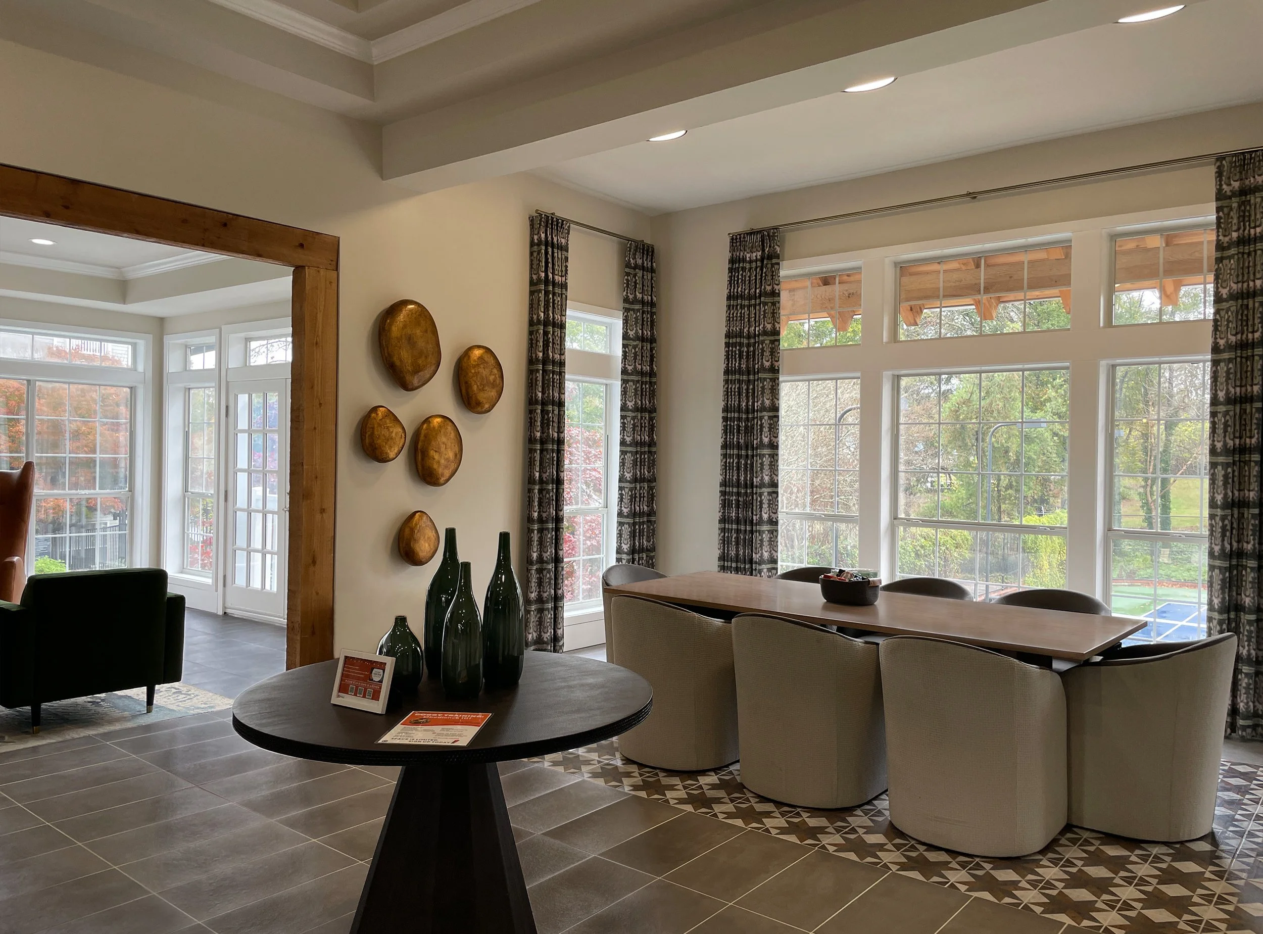 Interior of a modern dining area in an apartment lobby with large windows, a wooden table with six chairs, decorative wall art, and a round side table with black vases.