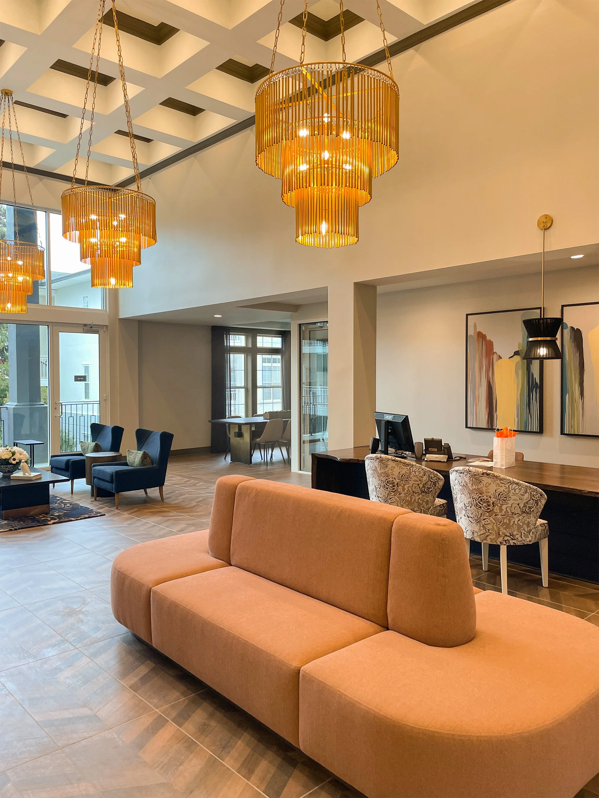 Modern hotel lobby with pink sofa, blue armchairs, a wooden desk with chairs, abstract wall art, large windows, and gold chandeliers.