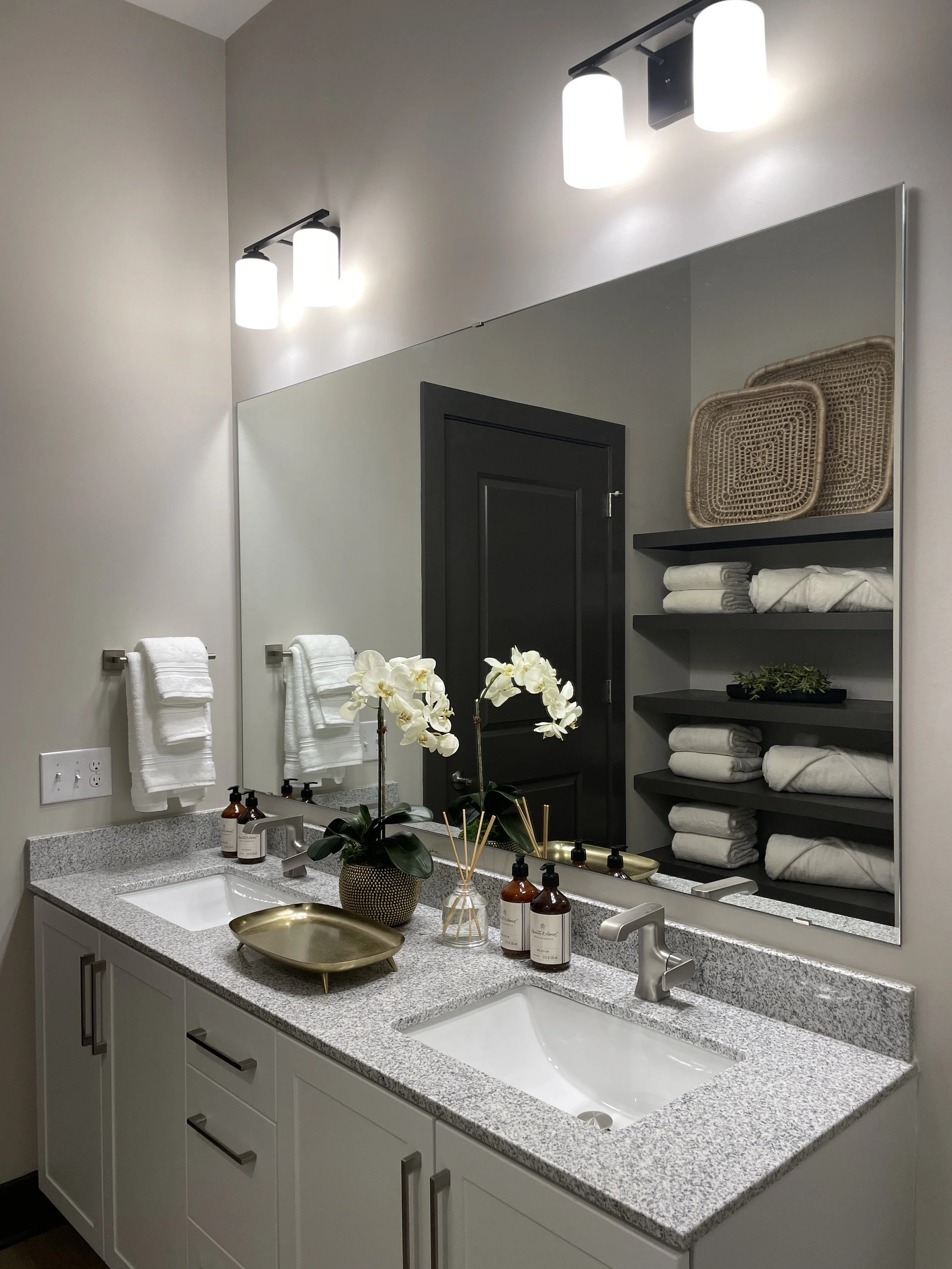 Bathroom vanity with a large mirror, two sinks, white cabinets, and decorative items including a orchid, soap dispensers, and a gold tray; towels are neatly folded on open black shelves, and a wicker tray is on the shelves above.