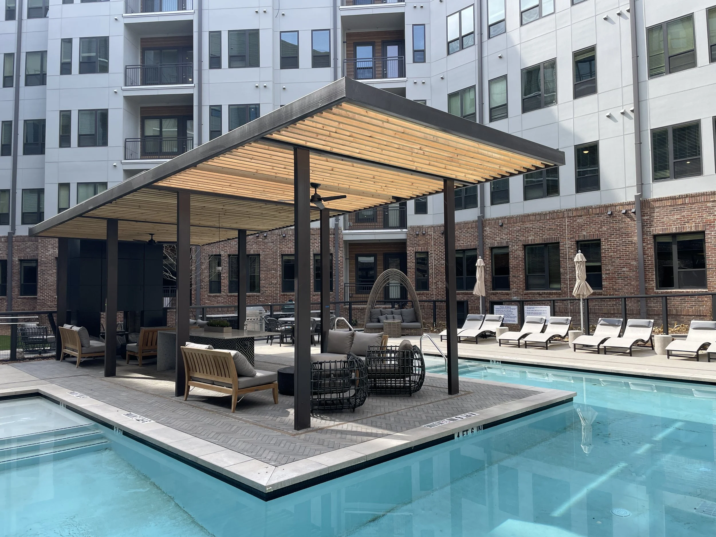 Swimming pool area of a modern apartment complex with lounge chairs, umbrellas, and a shaded seating area.