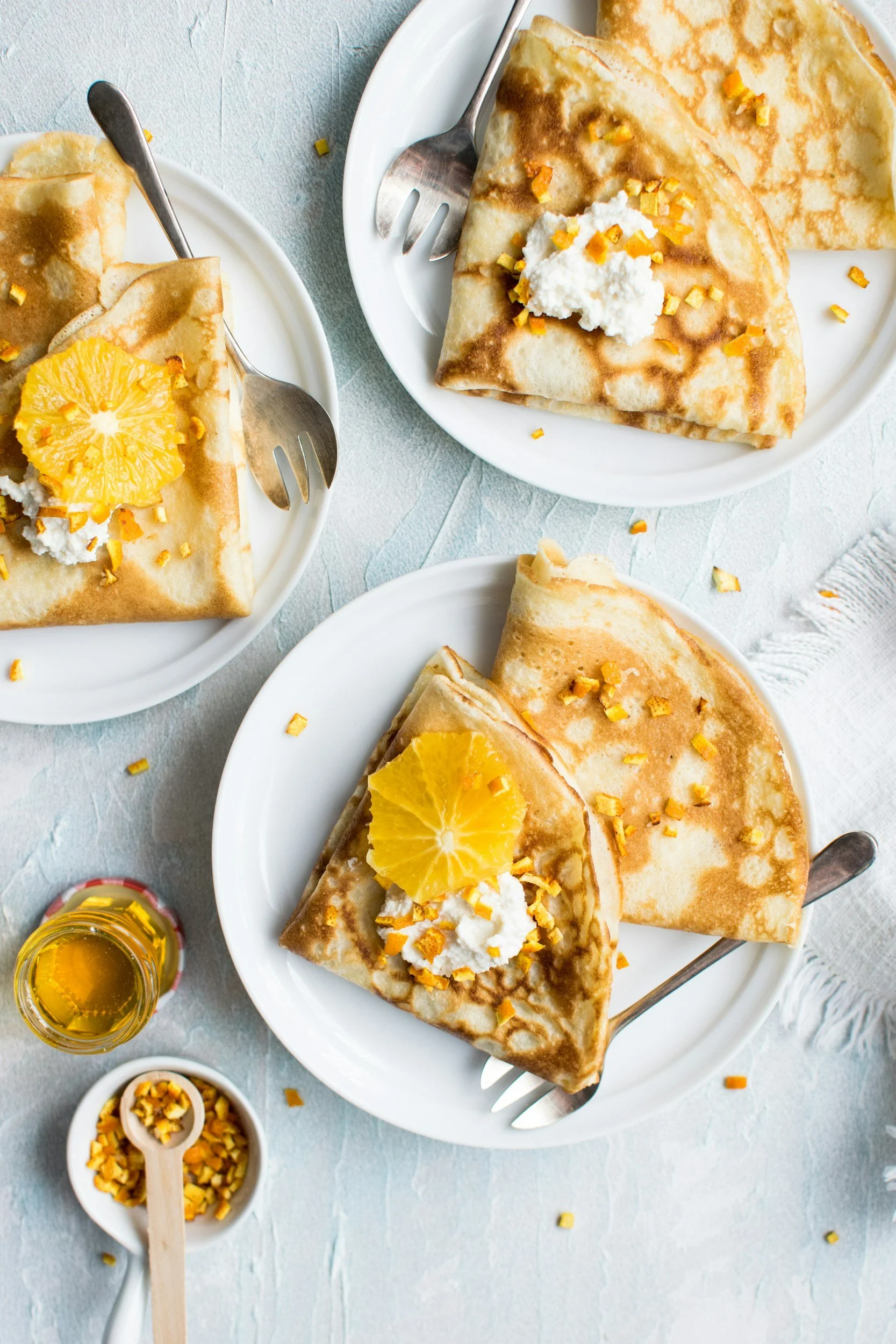 Three white plates each with a folded crepe topped with whipped cream, orange slices, and chopped nuts on a light surface, with a small bowl of chopped nuts, a small jar of honey, and a fork on each plate.