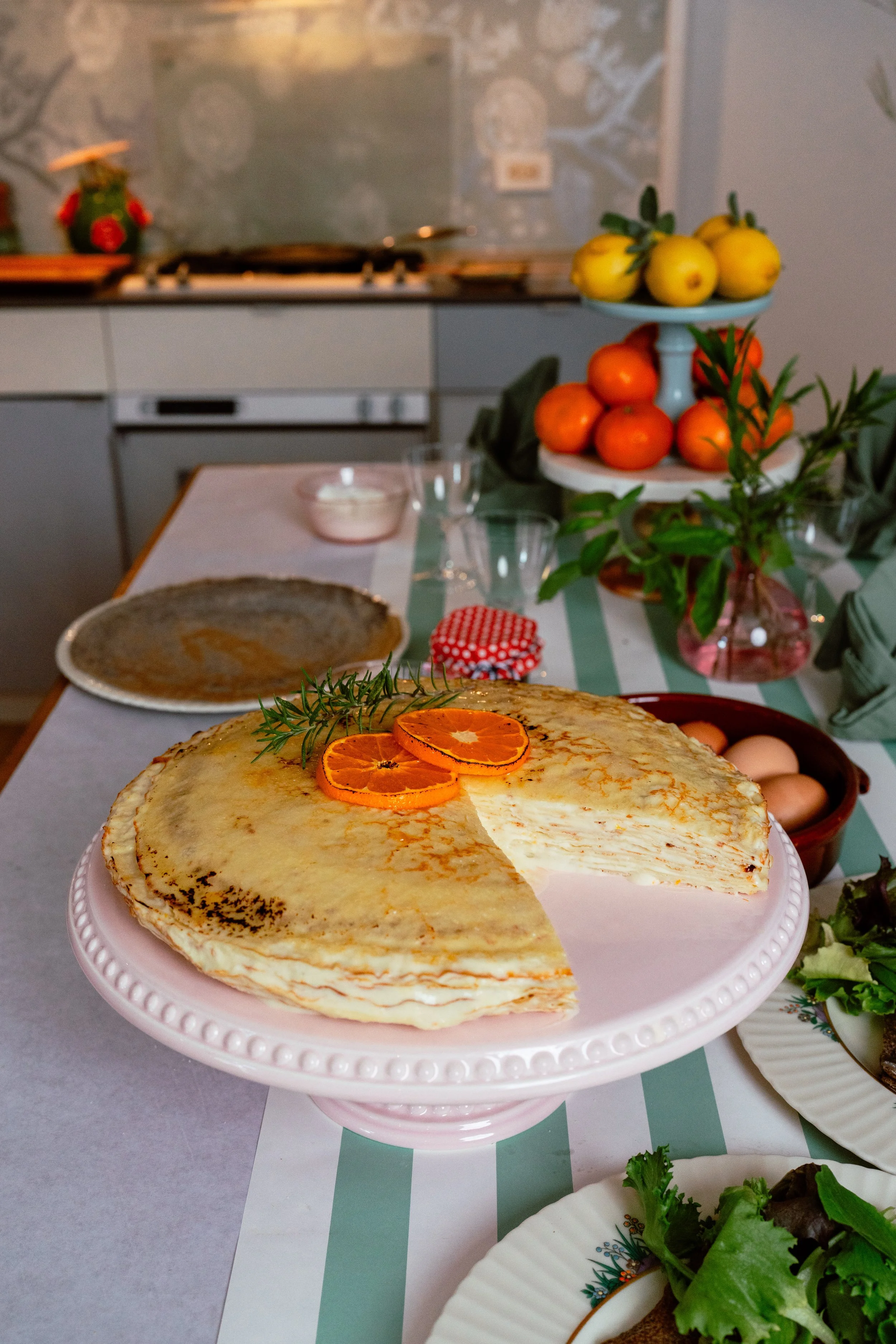 A crepe cake with slices of orange on top, garnished with a sprig of rosemary, placed on a pink cake stand on a dining table set for a meal.