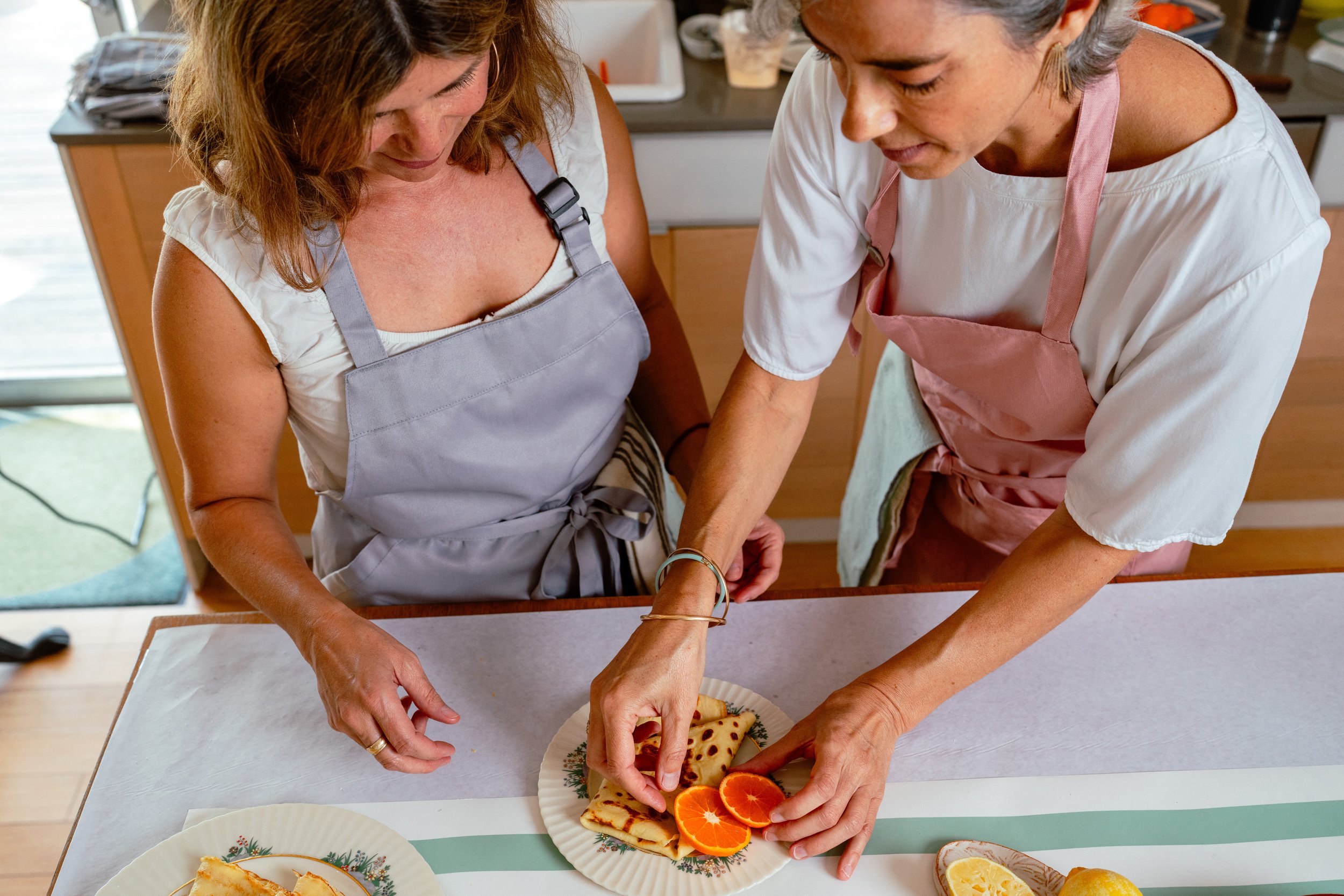Two women in aprons preparing food together in a kitchen, arranging sliced oranges and other food on a plate.
