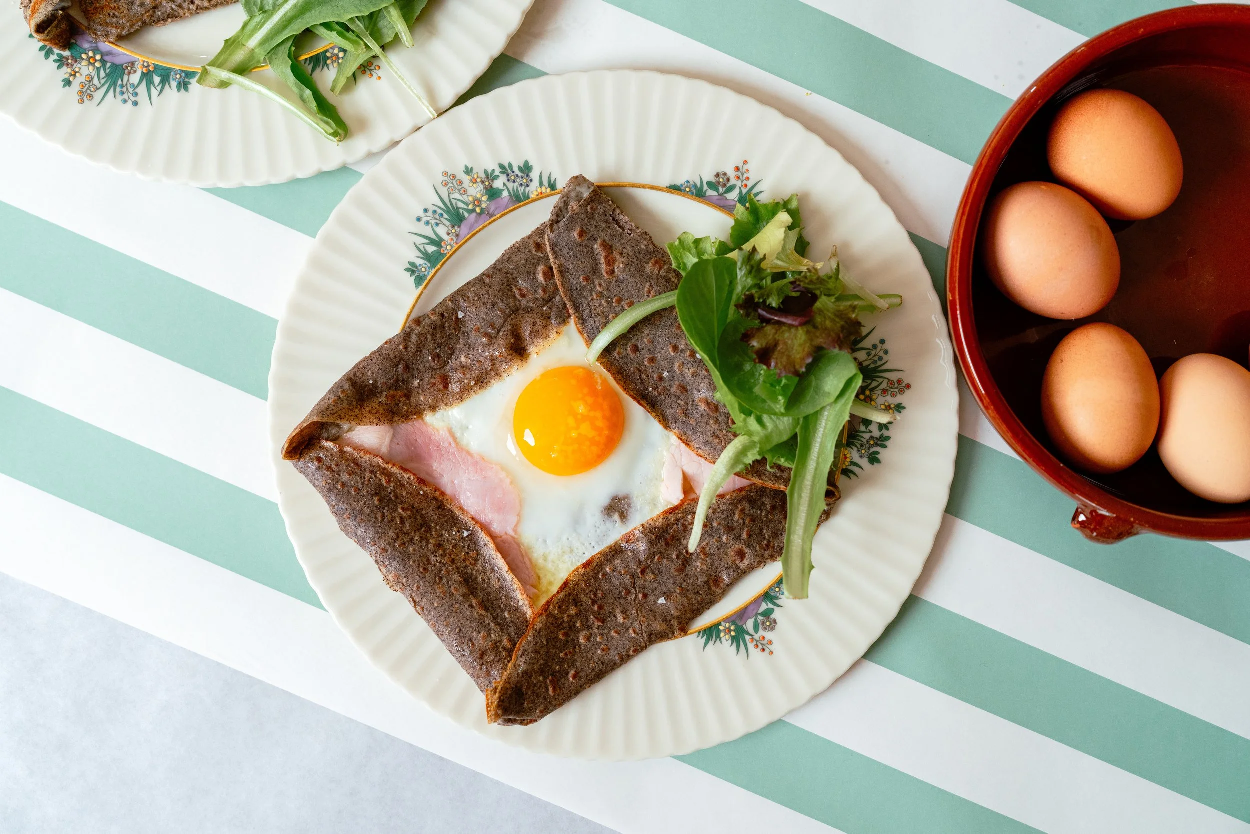 A plate with a buckwheat crepe, a fried egg, and salad greens, with a bowl of eggs nearby.